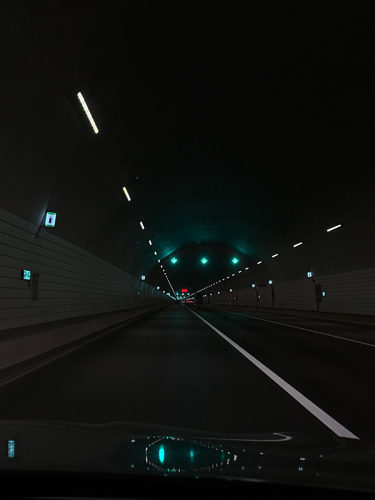 The image shows the interior of a tunnel, likely a road tunnel, with a dark ceiling and white tiled walls. Fluorescent lights are installed along the ceiling, emitting a cool white light, and some are angled downwards. Green directional arrows are illuminated on the ceiling in the distance, indicating traffic flow. The road surface is dark asphalt with a prominent white line marking the lane. The perspective is from a vehicle's dashboard looking forward through the windshield. In the foreground, the dark dashboard and windshield are visible, with reflections of lights from within the tunnel. On the left wall, there are several illuminated square signs, some with green borders and white symbols. The overall impression is of a modern, well-lit tunnel designed for vehicular traffic. This tunnel is located in Yiwu, China, likely part of the city's infrastructure.