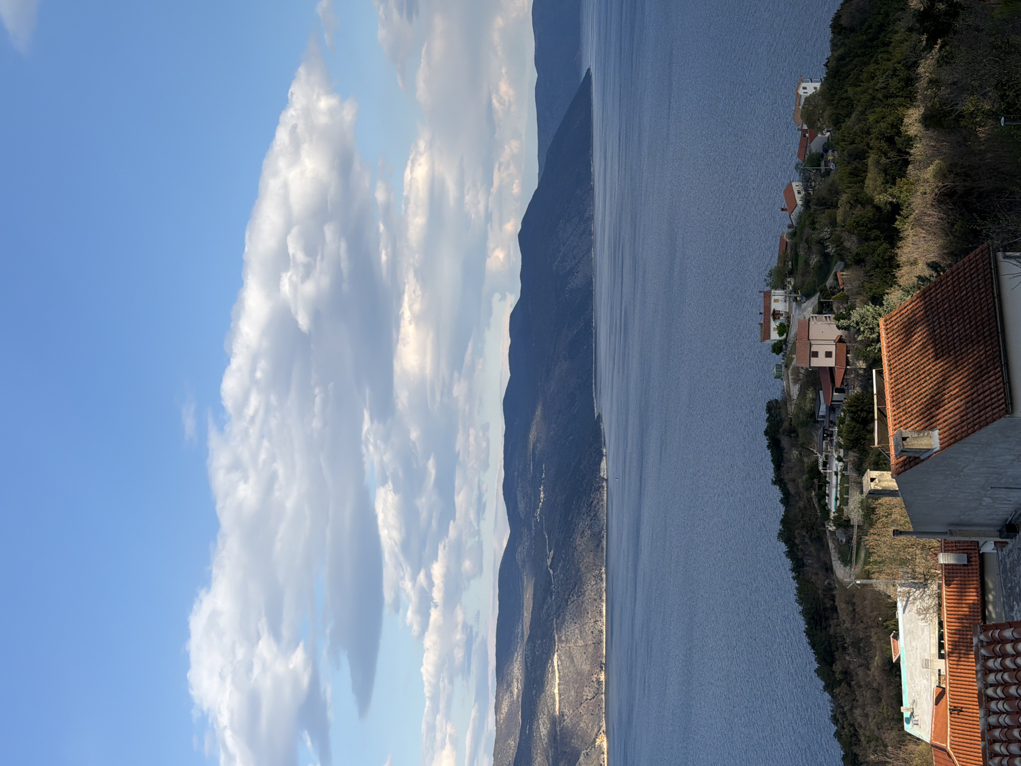 The scene depicts a view of Zagorje, Croatia, with a bright blue sky and fluffy white clouds on the left side of the frame. Moving rightward, a rugged mountain range with varying shades of green and brown flanks a body of water, likely the Adriatic Sea. The right side of the image showcases a coastal landscape with houses clustered along the hillside, intermingled with trees and vegetation. Some rooftops are visible in the foreground, suggesting the vantage point is from a higher elevation.