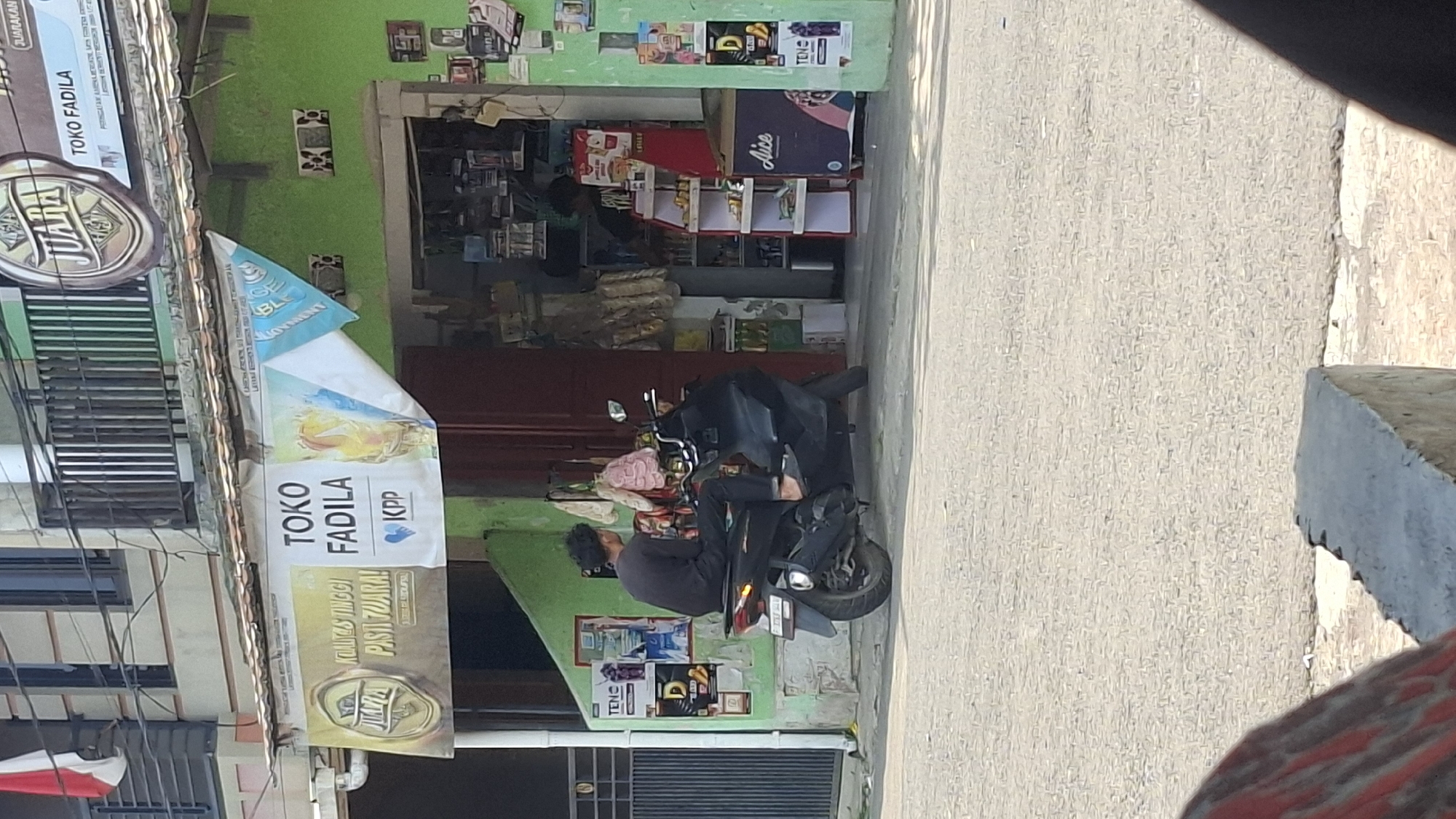 The image depicts a street scene in Cilaku, Indonesia, in front of a shop labeled "TOKO FADILA". A dark-colored scooter is parked on the asphalt road, with a young man wearing a black t-shirt and dark pants sitting on it, facing away from the camera. He appears to be barefoot. The shop, painted green, has its entrance open, revealing shelves stocked with various goods. A banner above the shop entrance displays "JUARA" and "TOKO FADILA" along with other text and graphics. To the left of the shop entrance, a poster is visible on the green wall, and a black metal gate is present further to the left. The ground in front of the shop and extending to the foreground is paved with a textured gray material, appearing to be a sidewalk or road surface. Sunlight casts shadows on the ground.
