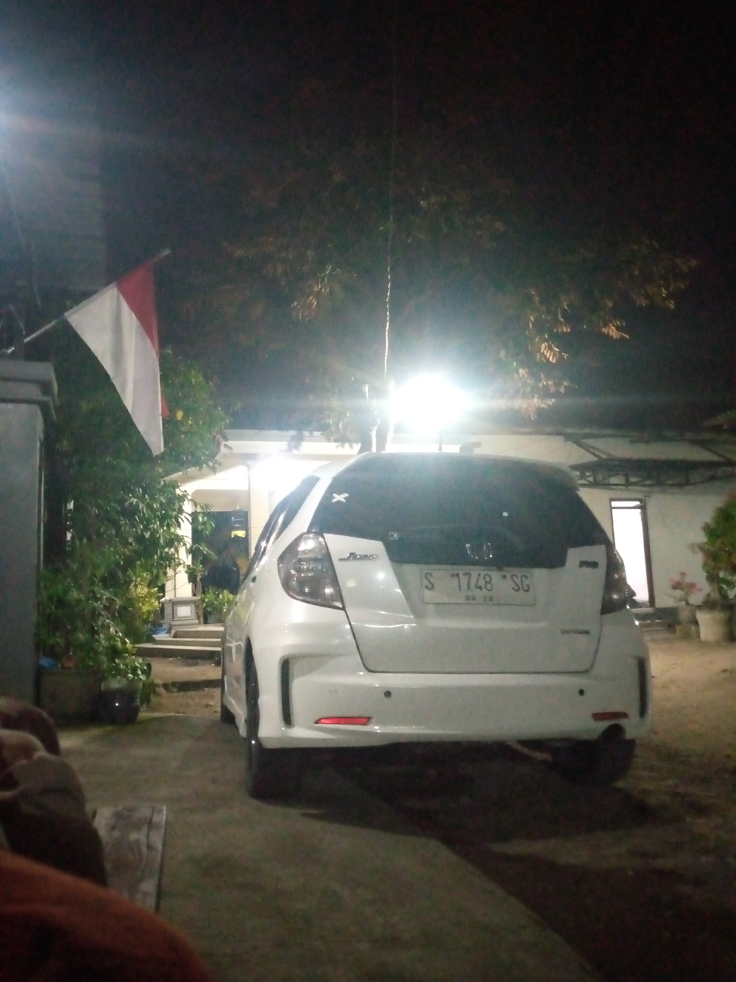 The image depicts a night scene in front of a building in Sumberwono, Indonesia. A white hatchback car is parked facing away from the viewer. The license plate is visible and reads "S 1748 SG". To the left of the car, there is a flag that appears to be the Indonesian flag, red and white, attached to a pole. Lush green foliage surrounds the flag and extends towards the building. The building itself has a porch with a light on and a doorway visible. The ground in front of the car and building is paved. In the foreground, a wooden bench is partially visible. The lighting suggests artificial illumination from the building and possibly streetlights. The overall atmosphere is dimly lit, characteristic of a nighttime setting. There are no people clearly visible in the image, though shadows suggest possible presence.