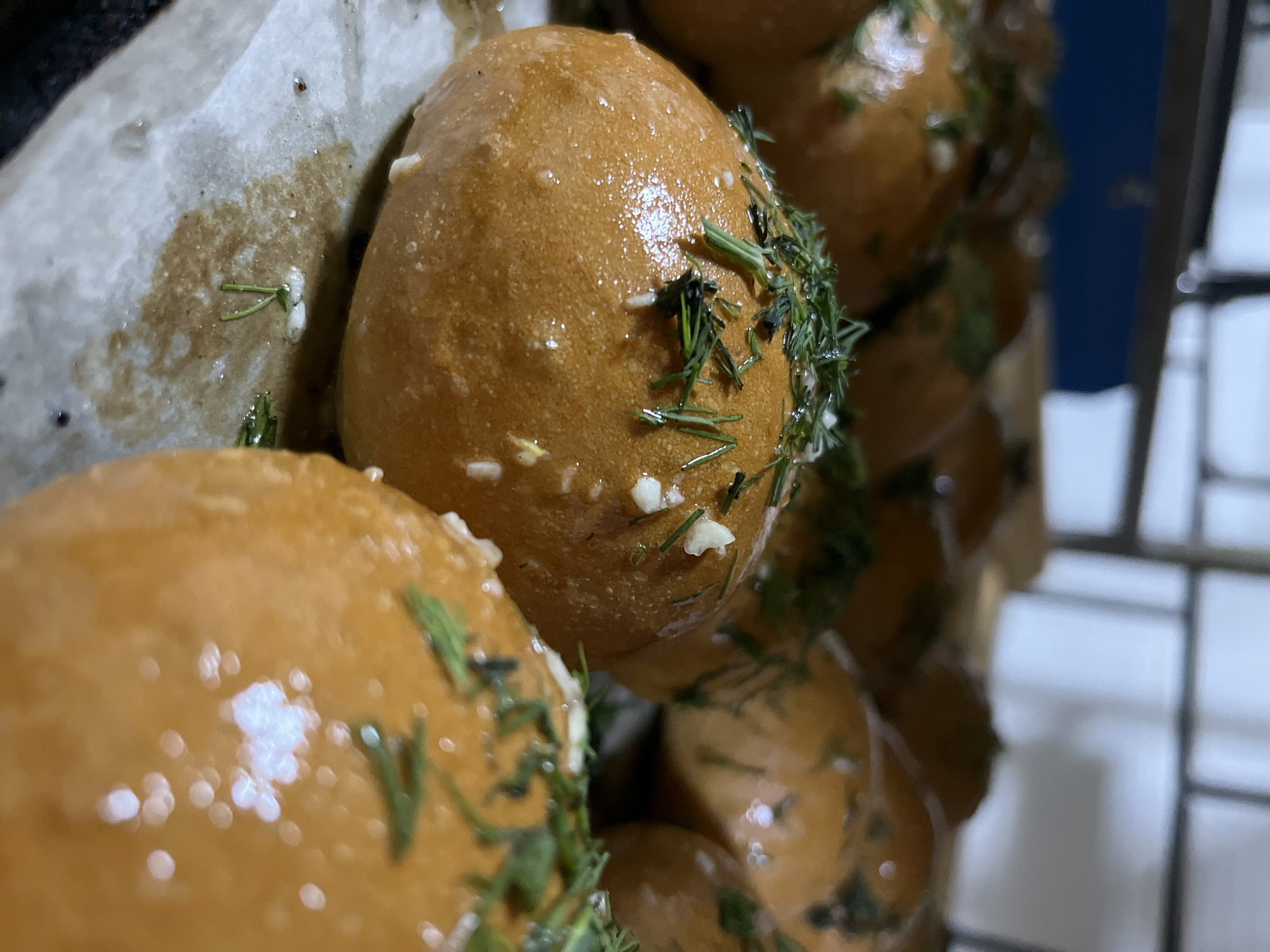 A close-up shot reveals a cluster of golden-brown buns, possibly freshly baked, arranged on parchment paper. The buns are liberally garnished with chopped dill and small pieces of what appears to be garlic. The glossy sheen on the buns suggests they have been brushed with oil or butter. The presence of parchment paper implies these buns might be part of a larger batch prepared in a domestic kitchen or small bakery. There's no specific evidence in the image that would locate this in Novopokrovskaya, Russia. The objects in the background appear to be part of a wire rack or shelf.