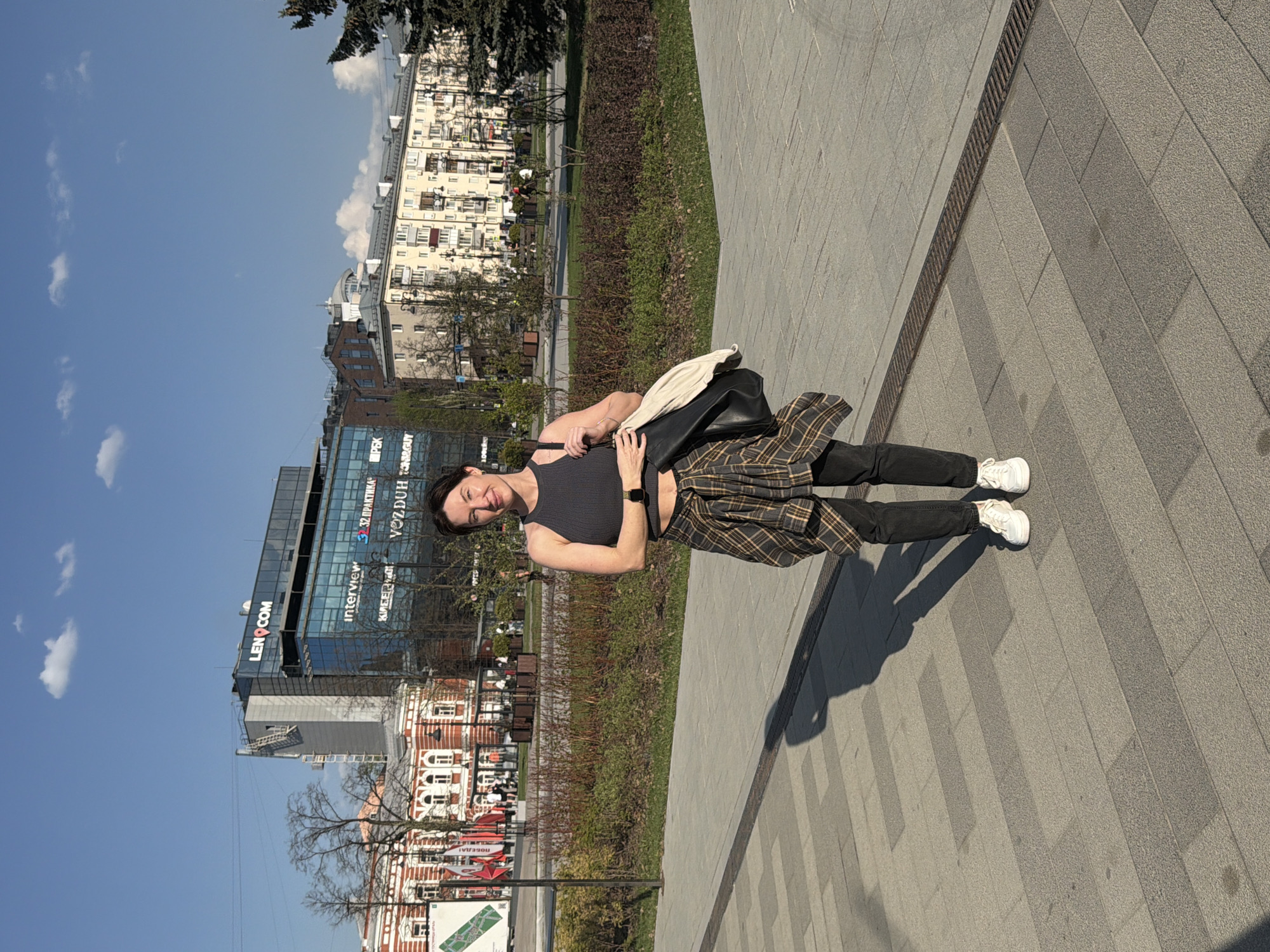 A medium shot captures a woman standing on a paved walkway in Perm, Russia. She is dressed in a gray tank top, black jeans, and white sneakers, with a plaid shirt tied around her waist. She carries a black bag with a light-colored cloth inside. The walkway is made of gray rectangular paving stones. Behind her is a green strip of lawn and a row of bushes, with buildings visible in the background. The buildings include a modern glass-fronted structure with "LENCOM" displayed prominently, as well as traditional brick buildings. The sky is blue with scattered clouds.