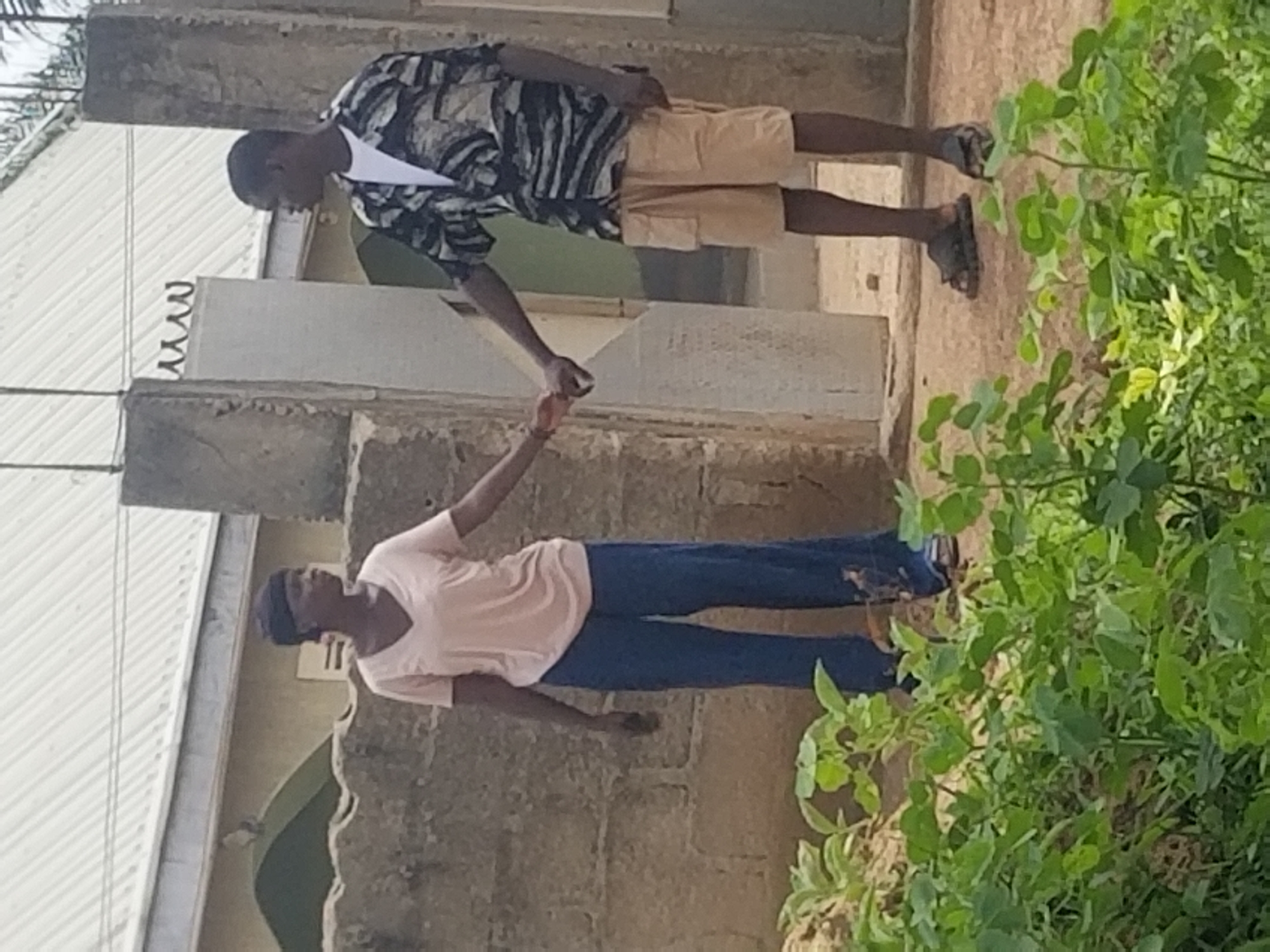 An outdoor medium shot captures two individuals, a woman and a man, standing near a stone wall and a gated entryway. The woman, on the left, wears a light pink v-neck t-shirt and dark blue wide-leg pants, with a dark head covering. She holds the hand of the man. The man, on the right, wears a white t-shirt, a dark patterned short-sleeved shirt, and light tan shorts. He is also wearing dark sandals. They are standing on a dirt path with green foliage in the foreground. In the background, a building with a light-colored corrugated metal roof and yellow walls is visible. A stone pillar and a section of a concrete wall with decorative metal spikes are part of the entryway.