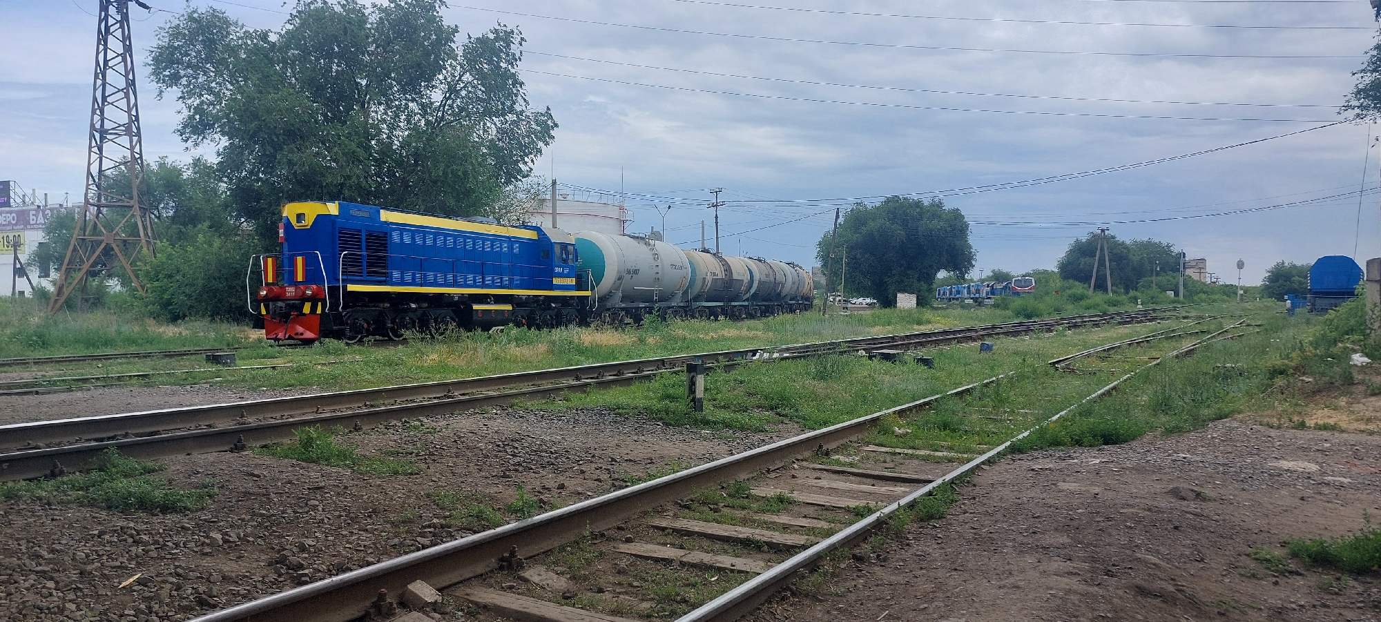 This image depicts a train yard on an overcast day in Oral, Kazakhstan. In the foreground, multiple sets of railway tracks run parallel to each other, with some overgrown with grass. In the middle ground, a blue and yellow diesel locomotive, identified as TE10-2419, is coupled to a series of gray tank cars. The tank cars appear to be empty. To the right of the tank cars, more railway tracks and a blue covered wagon are visible. In the background, trees and a power pylon frame the scene, with distant buildings and more trains on other tracks. The overall impression is of a functional, yet somewhat unkempt, railway facility.