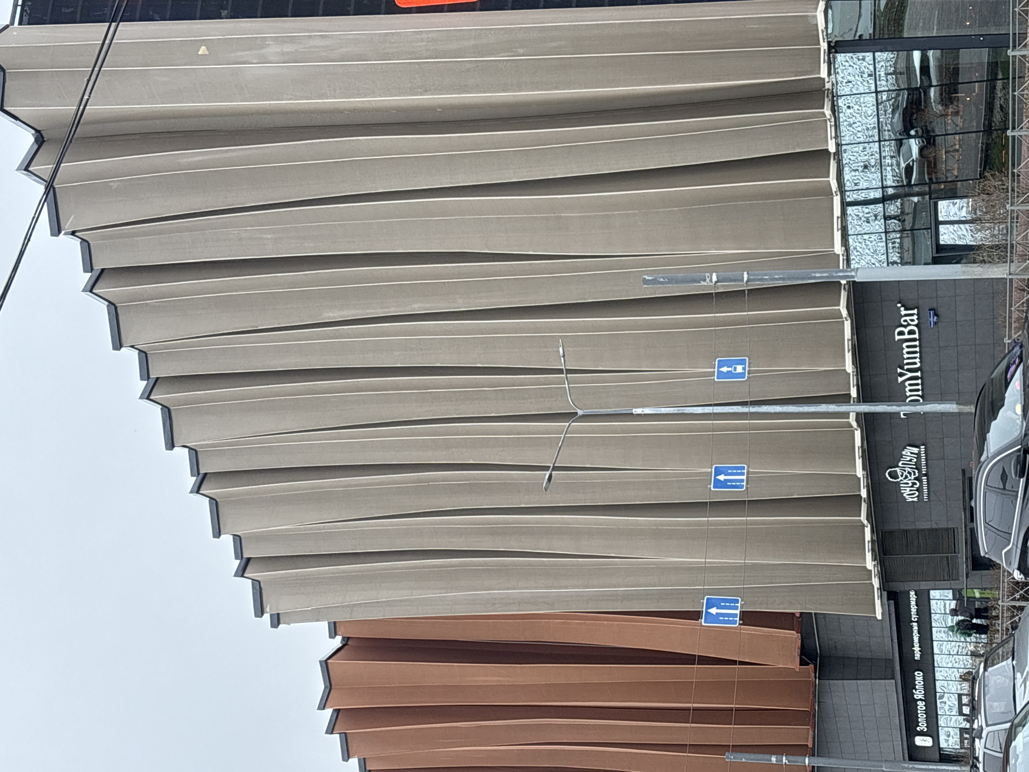 This media file captures an urban street scene featuring a modern building with a distinctive facade under an overcast sky. The building occupies the majority of the frame and is characterized by its layered, horizontally corrugated panels, creating a wavy, pleated effect. The upper section of the facade is light beige or grey, while the lower section transitions to a warm brown or copper hue. Black trim outlines the edges of these panels, adding definition to the architectural design.
On the ground level of the building, several commercial establishments are visible. Towards the right, a sign for "YumYumBar" is prominently displayed. Below it and to the left, another sign reads "КООП ПУРИ" (Koop Puri), identified as a "ГРУЗИНСКИЙ РЕСТОРАН" (Georgian Restaurant). Further left, the text "Золотое Яблоко" (Zolotoye Yabloko - Golden Apple) is visible, indicating a "парфюмерный супермаркете" (perfumery supermarket). The ground floor features large glass windows, some reflecting the street and vehicles.
Several street light poles with blue rectangular road signs displaying white arrows pointing straight are present, suggesting a road alongside the building. Cars are parked and potentially driving on the street in front of the building. In one of the cropped images, two small figures, presumably people, are seen near an entrance. The sky is uniformly grey and overcast, indicating daytime but without direct sunlight, contributing to a cool or muted lighting condition. Power lines or cables are visible extending across the upper left portion of the image. The Russian text on the signs confirms the location context of Perm, Russia.