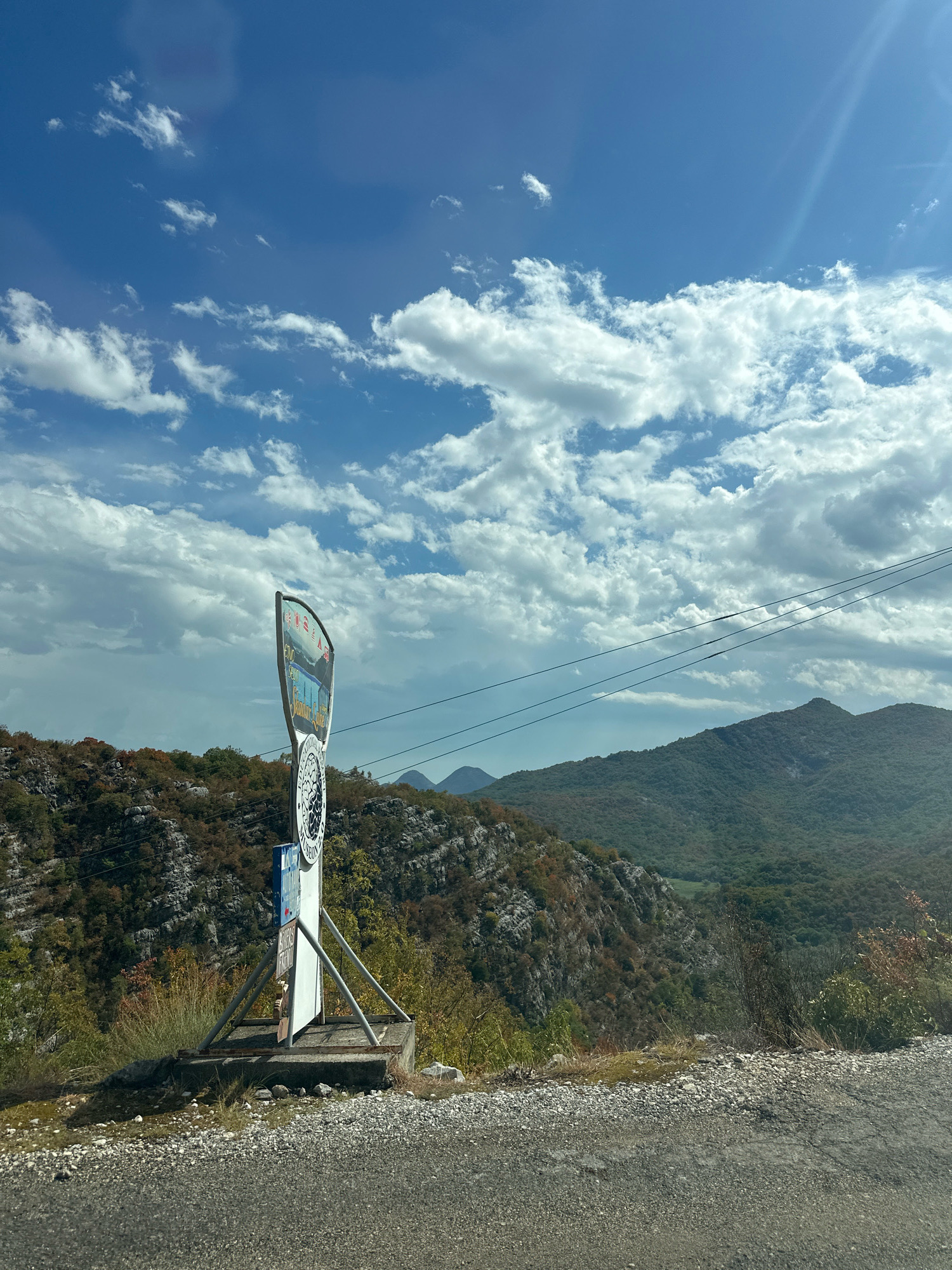 An eye-level, medium shot captures a clear blue sky with scattered white clouds, above a mountainous landscape in Cetinje, Montenegro. In the foreground, a grey asphalt road is visible, transitioning to a gravelly shoulder. A signpost stands on a concrete base, supported by metal legs. The sign itself is curved, featuring text and imagery, including what appears to be a circular emblem with a black and white design. Behind the signpost, the terrain slopes downwards into a valley filled with lush green trees. Rocky cliffs, partially covered in vegetation, are visible on the left side of the frame. In the distance, several green, tree-covered mountains form the horizon, with a hazy atmosphere. Power lines are strung across the middle ground. The overall scene depicts a scenic outdoor environment with a clear sky and mountainous terrain.