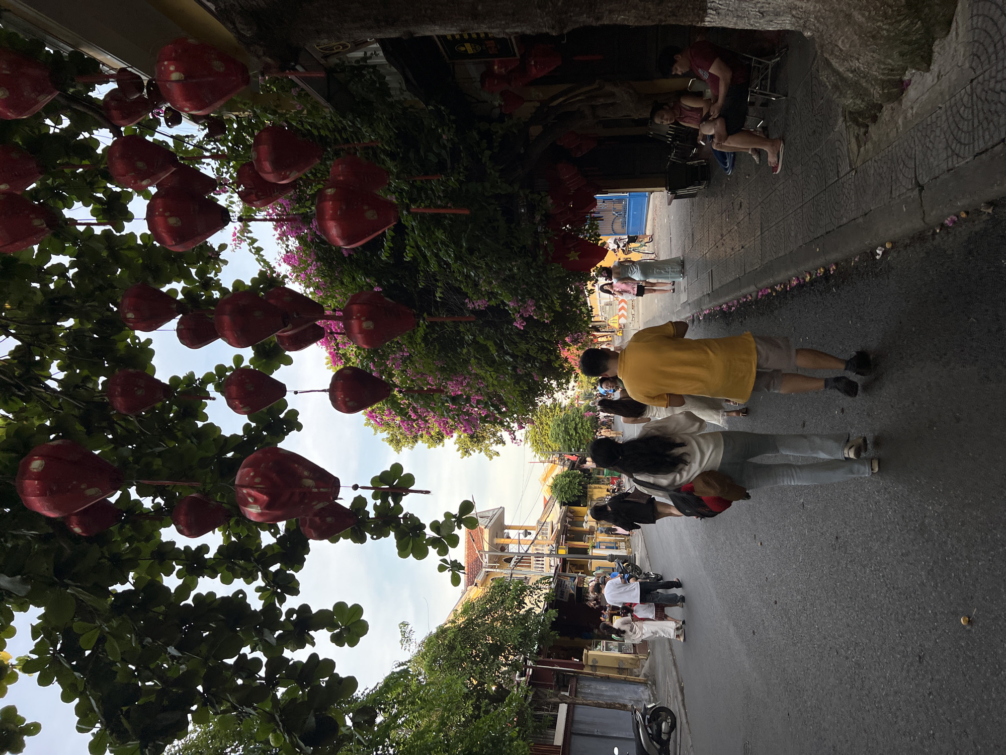 The image captures a street scene in Hoi An, Vietnam, likely during the late afternoon or early evening. Red, lantern-like decorations are suspended from the branches of leafy trees that overhang the street, casting a warm glow and creating a festive atmosphere. In the foreground, a couple walks away from the camera, their backs to the viewer. The woman wears a white top and light-colored jeans, while the man is dressed in a yellow t-shirt and shorts. Other pedestrians are visible further down the street, some walking, others sitting at outdoor establishments along the sidewalk. The architecture of the buildings lining the street appears traditional, with yellow facades and tiled roofs, characteristic of Hoi An's ancient town. A scooter is parked on the left side of the street. The overall impression is one of a bustling yet relaxed urban environment, enhanced by the decorative lanterns and the presence of people enjoying the evening.
