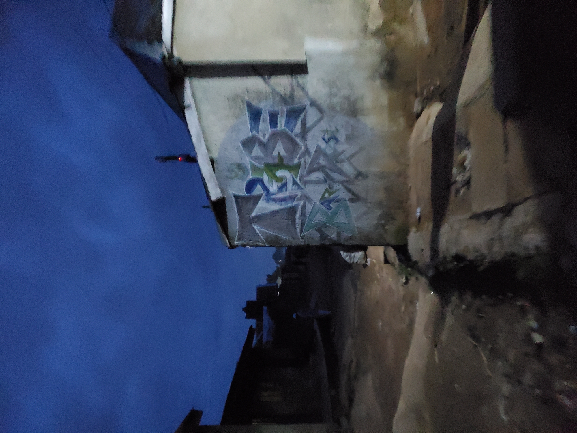 The image depicts a narrow alleyway in Jos, Nigeria, at dusk or night. The sky is deep blue. The alley is flanked by buildings, one of which has graffiti on its wall. The alleyway surface is rough and uneven. A single person is visible in the distance within the alley, though their features are indistinct. The light is dim, suggesting either the time of day or poor lighting conditions.