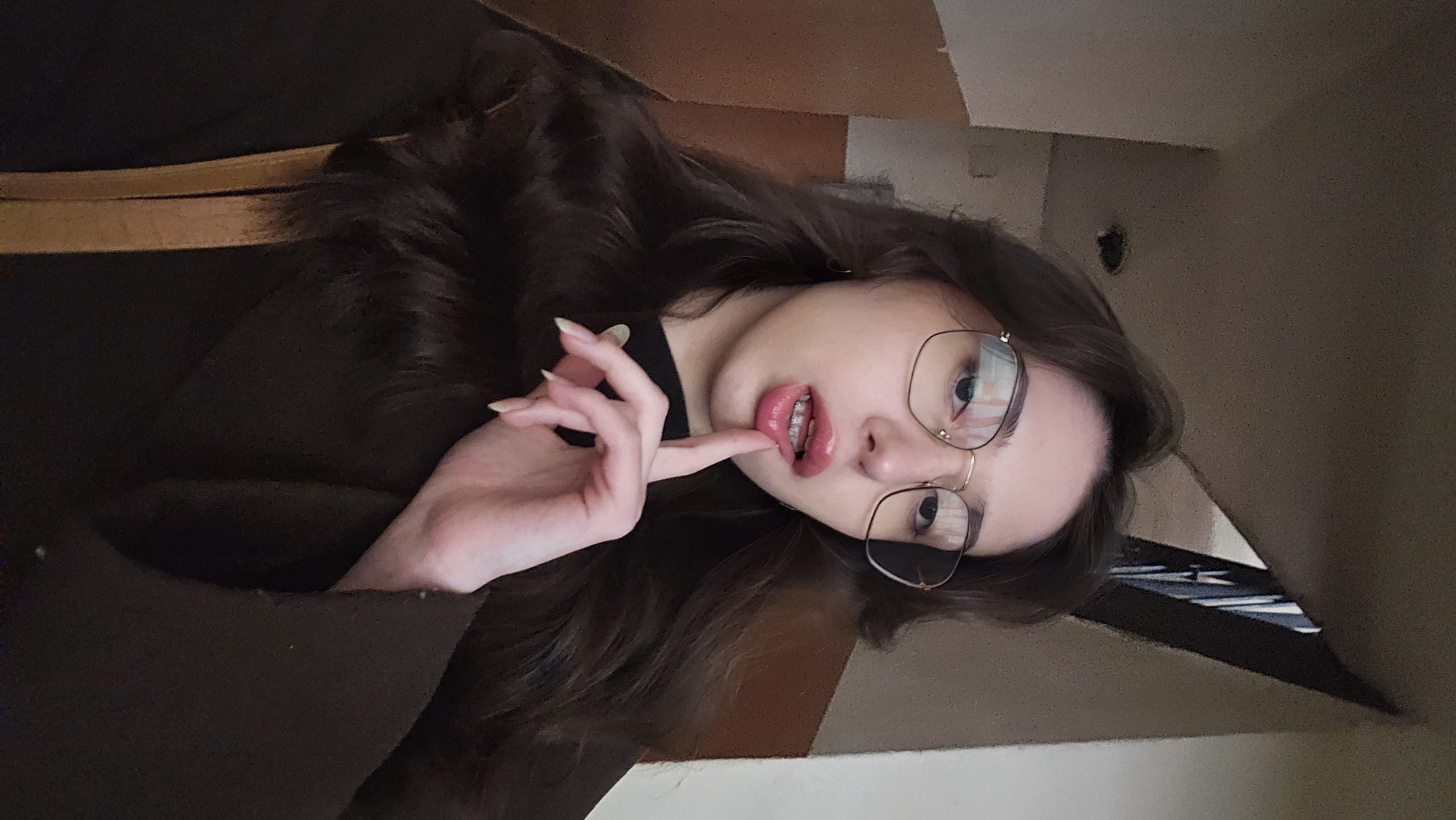A young woman with long brown hair, wearing glasses and dark clothing, is positioned in the foreground. Her left hand is raised near her mouth, with her index finger touching her lips. She appears to be indoors. The background includes a light-colored wall and a glimpse of what seems to be a dark object, possibly a vent or decorative piece. There is no visible evidence to determine the specific location in the city of Stara Zagora, Bulgaria.