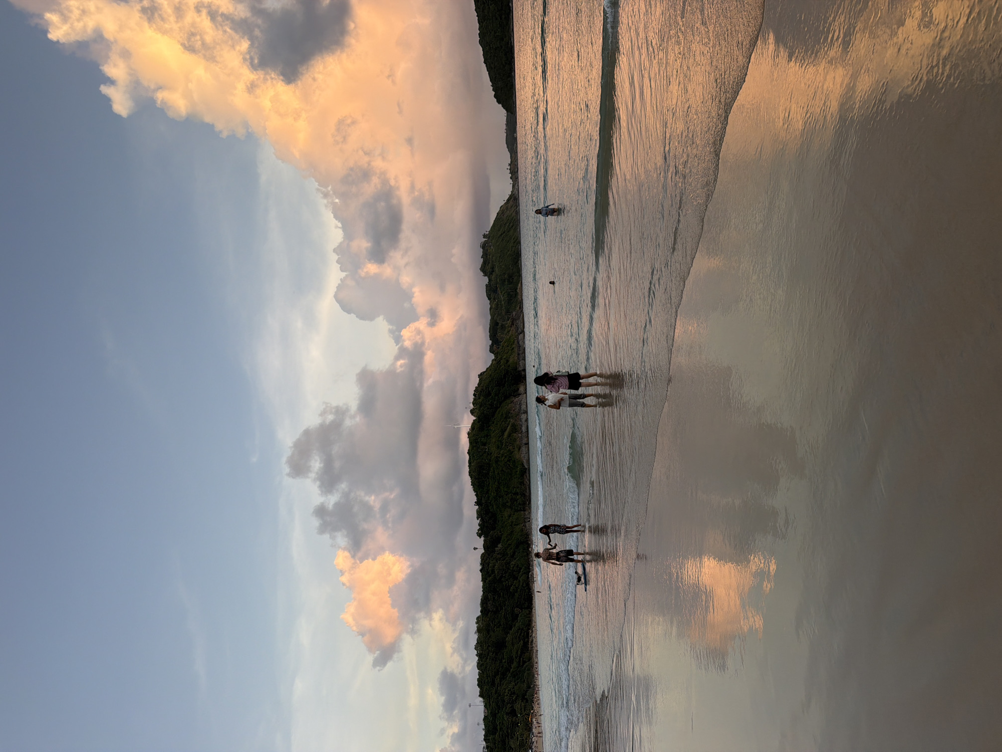The image depicts a serene beach scene at sunset in Rawai, Thailand. The sky is a gradient of soft blues and pinks, with dramatic, illuminated clouds painting the horizon. The gentle waves of the Andaman Sea lap at the shore, creating a reflective surface on the wet sand, mirroring the colors of the sky and clouds. In the foreground, a few individuals are visible wading in the shallow water. A couple stands hand-in-hand, facing the sea, with what appears to be a small surfboard or paddleboard at their feet. Further out, other people are scattered, some swimming or standing in the water. The background features a lush, tree-covered hillside, characteristic of the Rawai coastline, with a few distant structures or lights visible. The overall impression is one of tranquility and natural beauty as the day draws to a close.