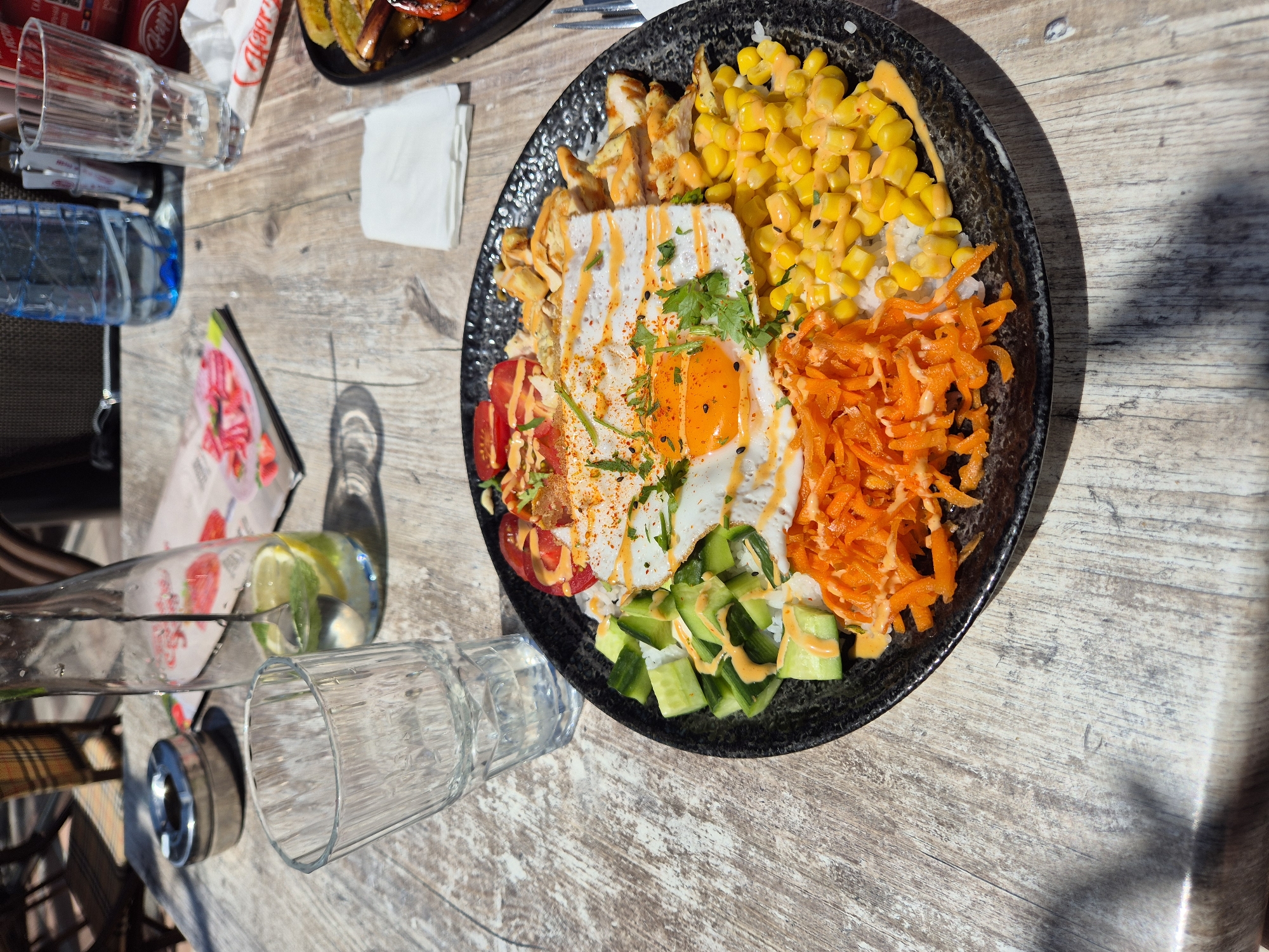 The image captures a top-down view of a table setting, likely in an outdoor cafe or restaurant in an unidentified location. A plate containing a colorful dish is the focal point. This dish includes rice, corn, shredded carrots, sliced cucumbers, tomatoes, and chicken. The dish is topped with a fried egg decorated with herbs and a sauce. Beside the plate, there are several drinking glasses, one of which is tipped over. A water bottle is present, along with a menu card. The table has a weathered, wooden surface. A second plate is barely visible in the top left corner.
