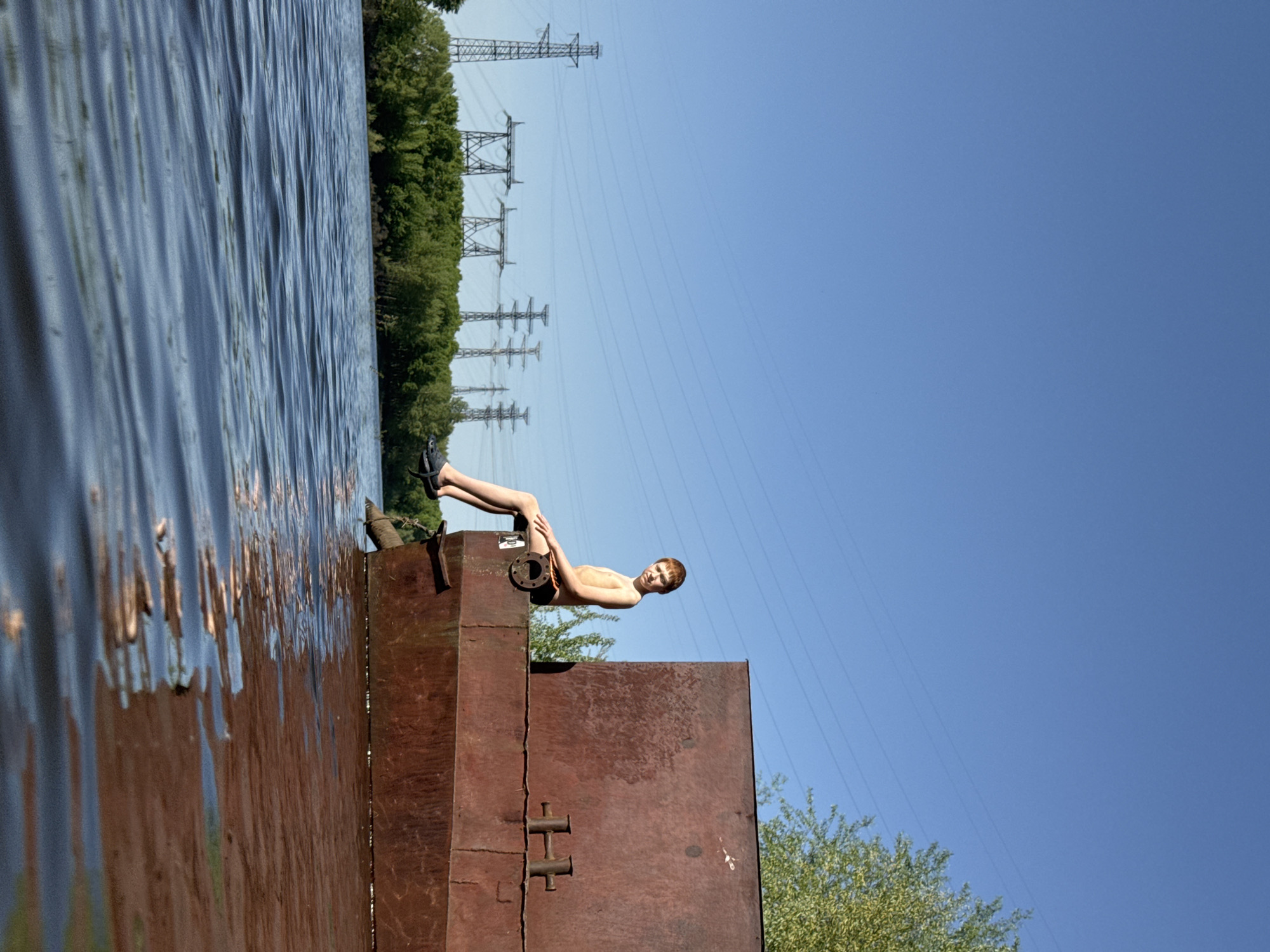 A boy is sitting on the edge of a metal structure beside water, seemingly in the city of Chaikovsky, Russia. The water is rippled and reflects the sky and surrounding foliage. Power lines and trees are visible in the background. The metal structure is rusted and has some metal fittings on its side. The boy is light-skinned and wearing dark shorts and shoes. He is leaning back and looking towards the viewer. The sky is blue and mostly clear.