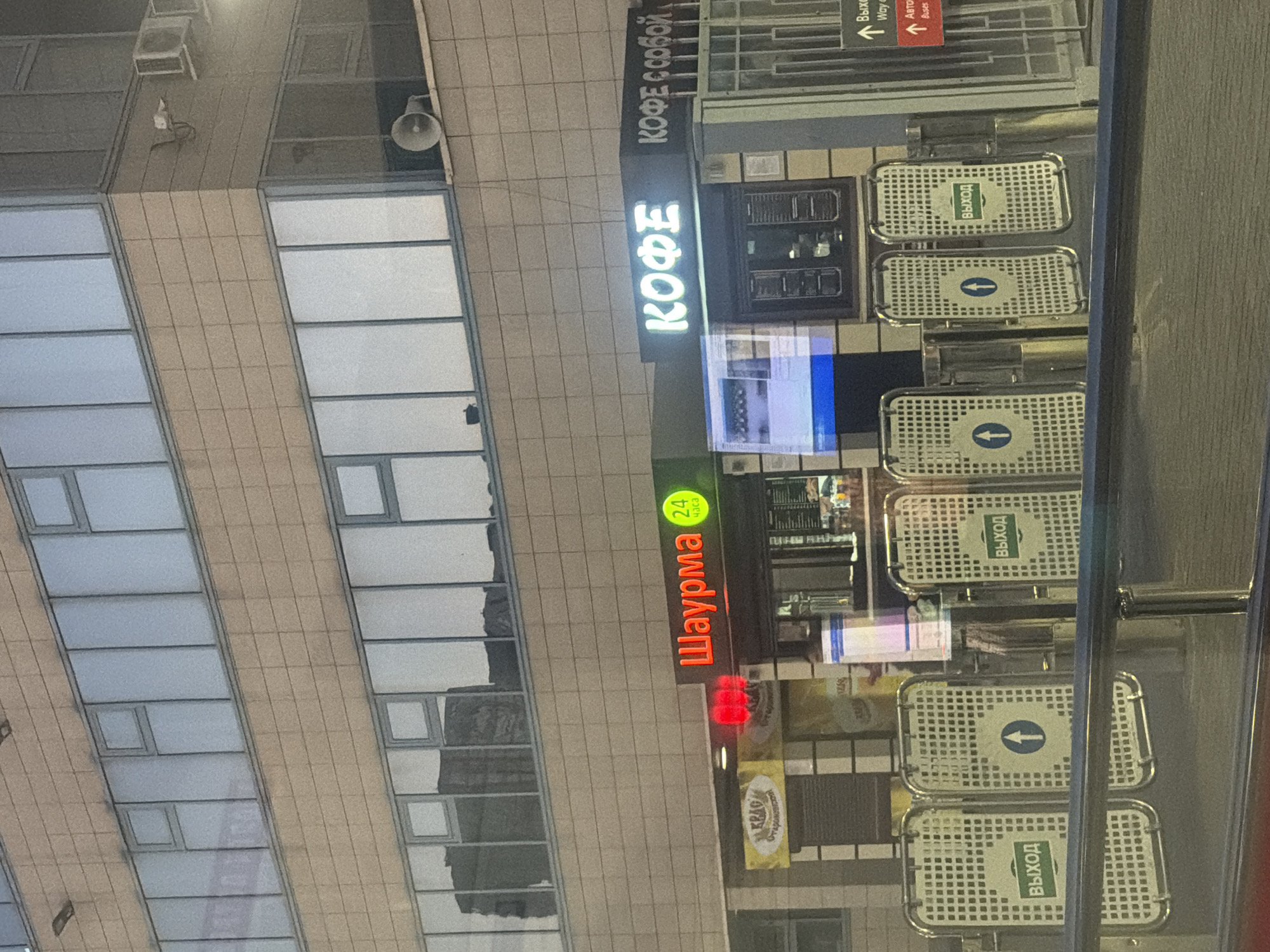 The image shows a street-level view of a modern building with several shops and entrances. On the left, there is a business with yellow signage that reads "KRAG" in stylized text, followed by Russian text. Adjacent to this is a shop with bright red neon lettering spelling "Шаурма" (Shawarma) with a green circle containing "24" and "часа" (hours) indicating it is open 24 hours. To the right of the shawarma shop is a neon sign with white lettering that says "КОФЕ" (Coffee), and to its right, smaller text reads "КОФЕ С СОБОЙ" (Coffee to go). In front of the shops, there are multiple sets of pedestrian turnstiles, some with green signs displaying the Russian word "ВЫХОД" (Exit) and an upward arrow symbol. One turnstile has a blue upward arrow symbol on a white circular background. Behind the turnstiles and shops, there is a glimpse of a bright blue illuminated screen. The building facade is constructed of light brown brick-like panels and features large, reflective windows. On the upper part of the building, a white horn speaker is visible mounted on the exterior wall. Signs indicating directions for "Way out", "Авто" (Cars), and "Buses" are also visible on the right side of the frame. The overall scene depicts a commercial area with food establishments and controlled pedestrian access.