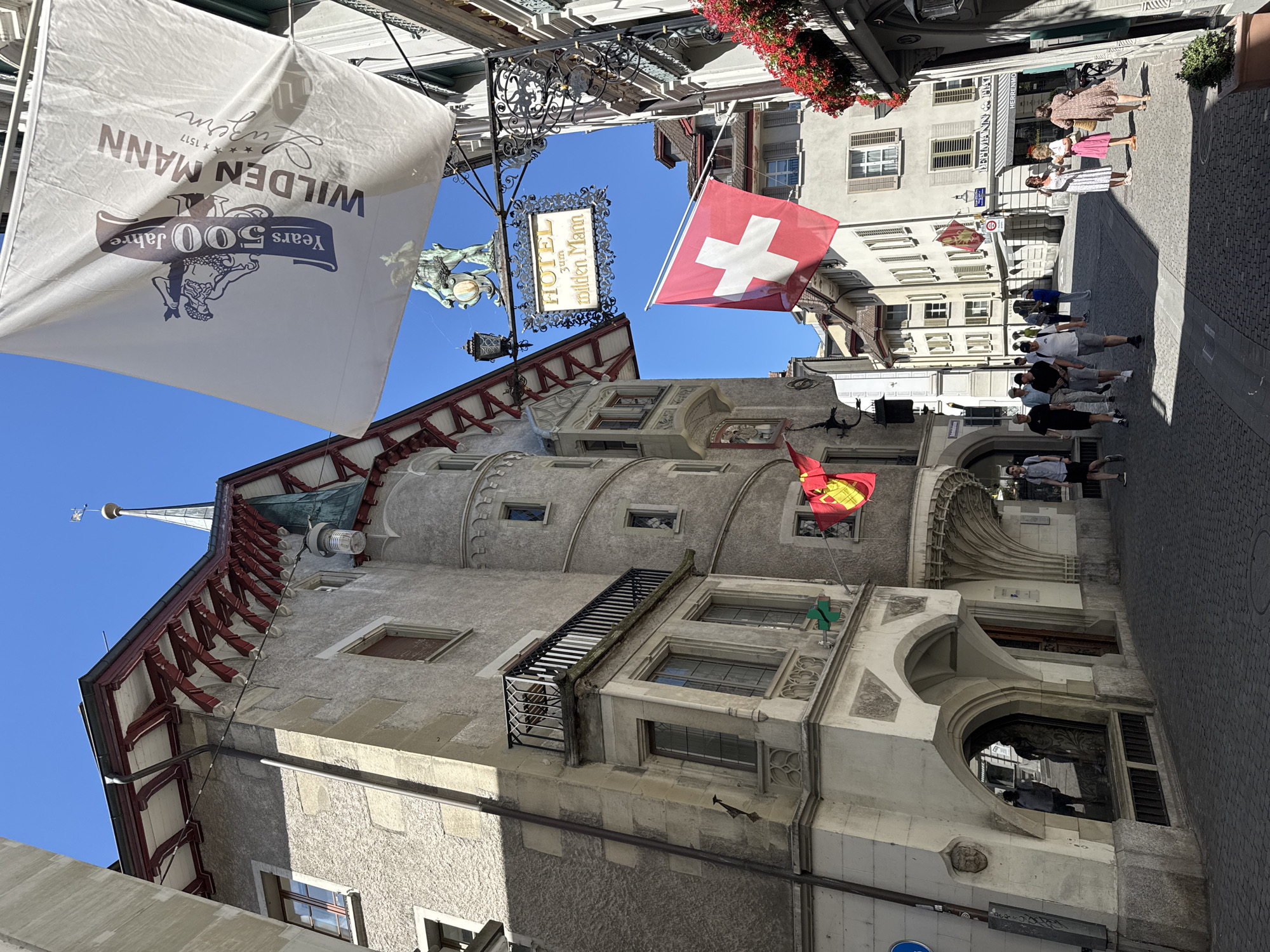 This image captures a sunlit street scene in Lucerne, Switzerland, specifically in the historic Old Town, likely near the Chapel Bridge or the Jesuit Church. Dominating the view is the striking architecture of the Hotel Wilden Mann, a landmark building with its distinctive turrets, gabled roof, and intricate stonework. A large white flag, commemorating "500 Jahre Wilden Mann Luzern," hangs prominently, advertising the hotel's long history.
Below the hotel's signage, a Swiss flag waves in the breeze, a clear indicator of the country. The street is paved with cobblestones, typical of European old towns, and lined with various buildings, some displaying flowers in window boxes. A diverse group of people are strolling through the street, some in casual attire, others dressed more formally, suggesting a mix of tourists and locals.
In the foreground, on the left, a unique building facade features arched entrances and detailed stone carvings. To the right, a sign for "Lehmann & Fiat" and "Herrenmoden" indicates shops or businesses. The overall atmosphere is lively and picturesque, characteristic of a vibrant European city on a clear, sunny day. The presence of the historic hotel, the Swiss flag, and the architectural style strongly points to Lucerne's Old Town as the location.