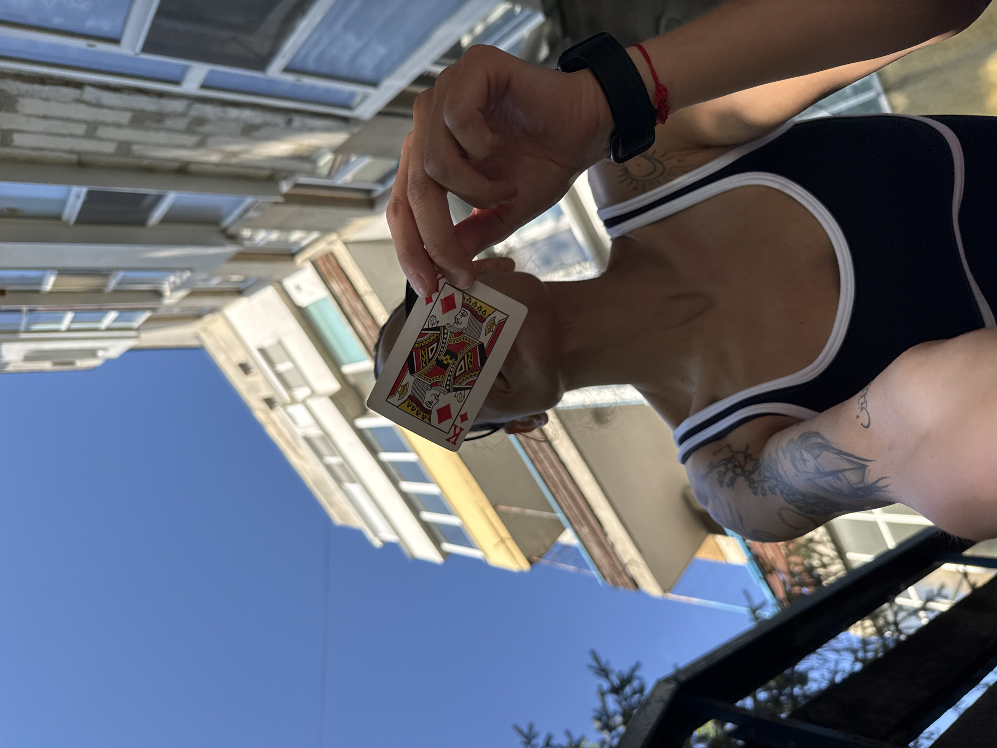 A person with tattoos is holding a King of Diamonds playing card in front of their face. The person is wearing a black and white tank top and a black watch on their wrist, which also has a red string tied around it. Buildings and a blue sky are in the background. Vegetation is also visible at the bottom of the frame, and a metal structure is visible near the bottom right corner. The image is taken from a low angle, angled upwards. While the exact location is not discernible, the description includes Pleven, Bulgaria as a potential location.