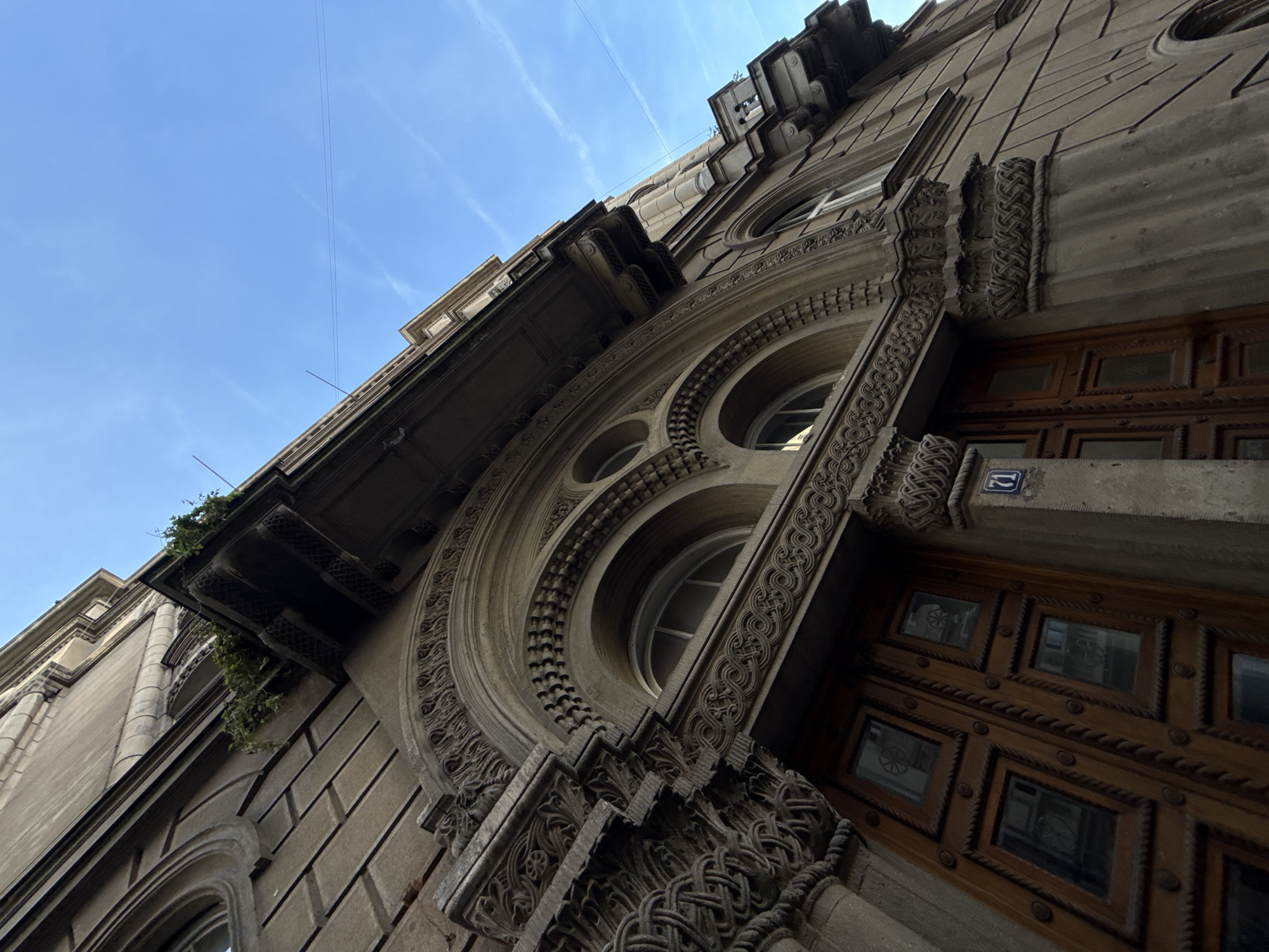 A low-angle shot depicts the ornate facade of a building in Belgrade, Serbia. The building's exterior features carved stonework around arched windows and doorways. A wooden door with a grid pattern and small window panes occupies the right side of the frame. A small, blue and white address plaque is mounted near the door. A balcony with greenery overhangs the facade. The sky above is clear blue.