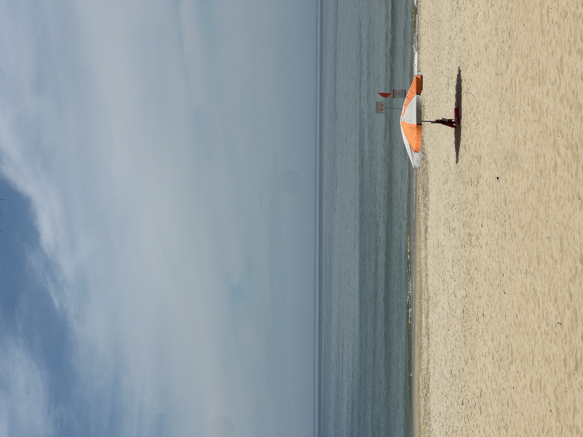 An orange and white beach umbrella stands on a sandy beach, casting a shadow. A person is seated under the umbrella. Two signs, one with red and white warnings and a red flag, are positioned behind the umbrella. The ocean stretches out to the horizon under a partly cloudy sky. The location is Unknown, Unknown.