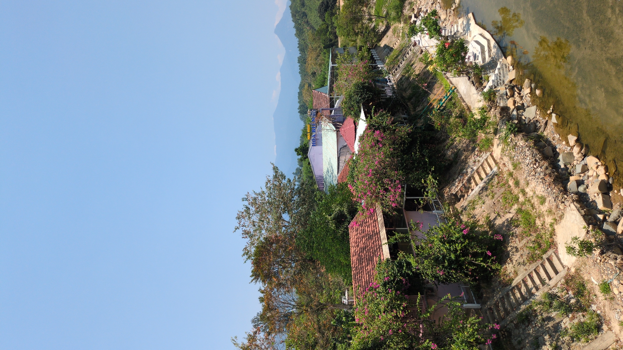 A landscape photograph depicts a lush, hillside view with a focus on architecture and natural elements. The sky, a clear azure blue, dominates the upper left portion of the frame. Below this, there is a verdant hillside with varied foliage, including trees with autumn-colored leaves and flowering bushes with pink blossoms.
Buildings with terracotta-tiled roofs and light-colored facades are nestled within the greenery. A restaurant called "Hoa Bien" with blue lettering on the roof can be spotted among the houses.
A rocky riverbank lines the right edge of the picture. A cascading set of concrete steps leads down to the water, providing access to the clear river. A scattering of stones and boulders can be seen both on the bank and partially submerged in the water.
The scene seems to be located in Nam Khánh Vĩnh, Vietnam, based on the presence of the restaurant. The photo appears to be taken from an elevated vantage point. There are no people or animals visible in the image. The overall composition suggests a peaceful, natural setting with integration of human-built structures.