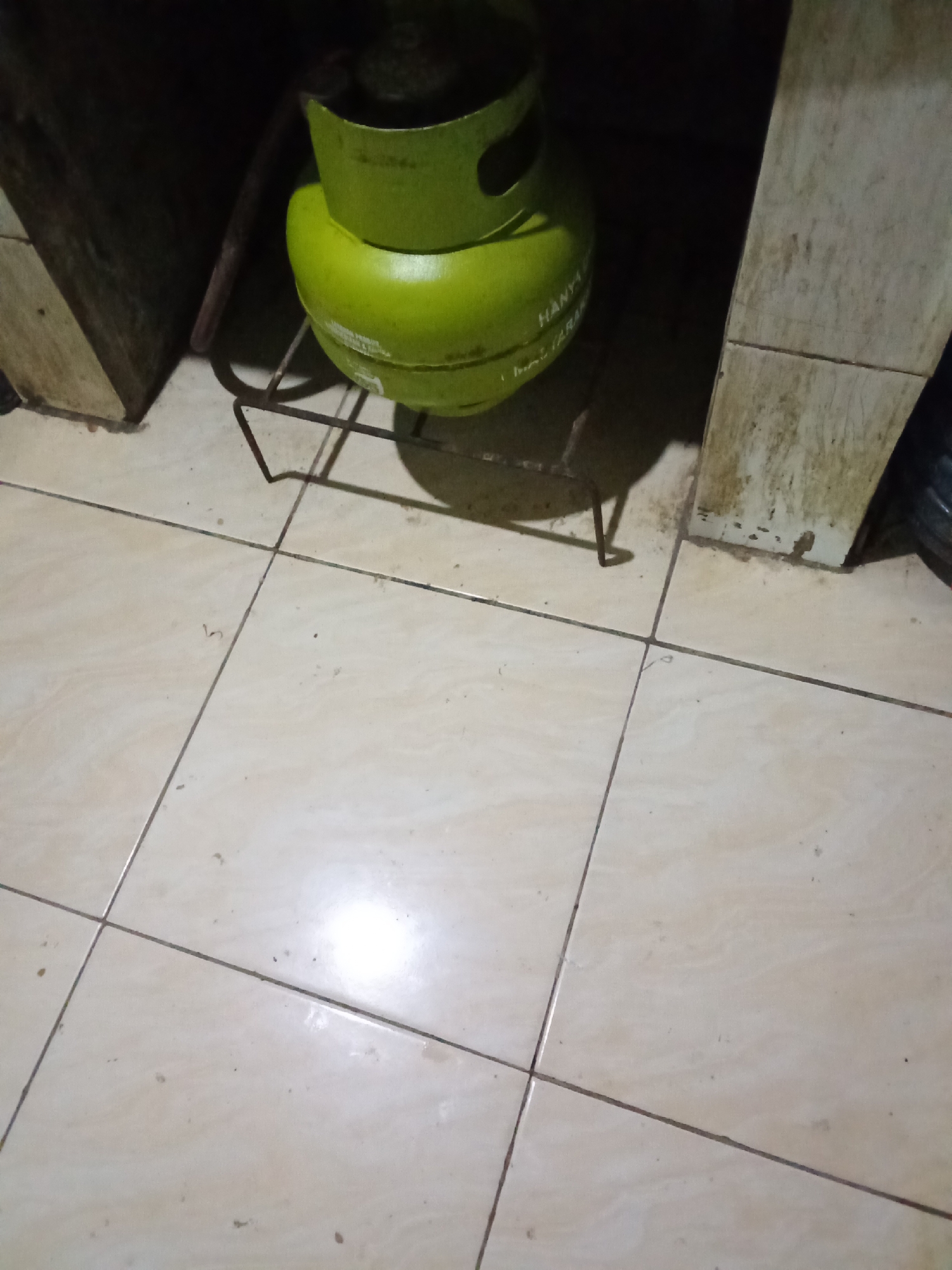 A green LPG gas cylinder rests on a metal stand on a tiled floor. The cylinder has text printed on it, which appears to be in Indonesian. The background is dark, with some light-colored wooden structures on the left and right. The overall environment suggests an indoor setting, possibly a kitchen or storage area in a home or business in Pemalang, Indonesia. The lighting is dim, with a strong light source casting shadows. There are no people or animals visible in the image. The floor tiles are a light beige color with dark grout lines. There are some minor stains and debris on the floor and on the wooden structures.