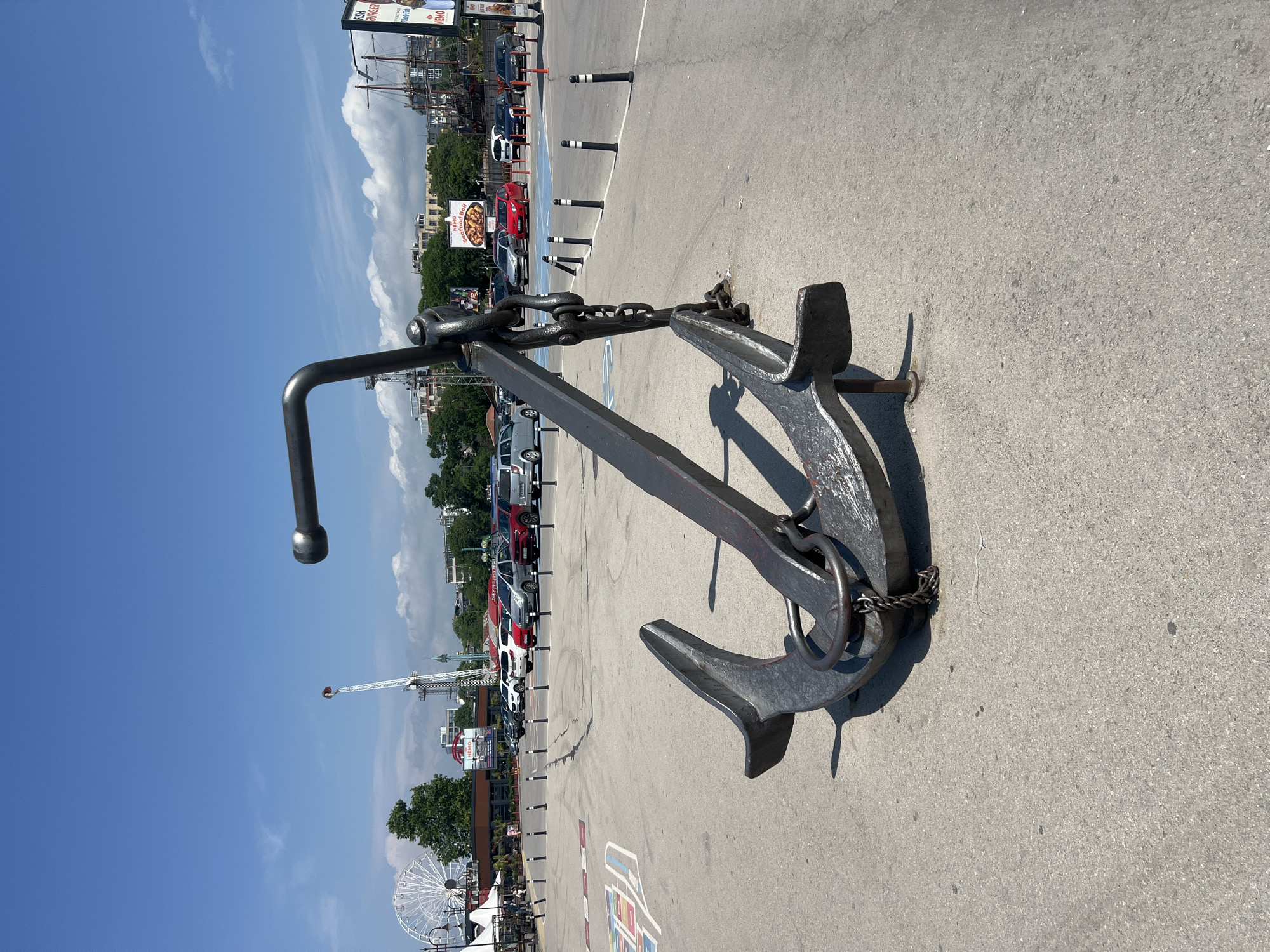 The image, rotated 90 degrees counter-clockwise, depicts a bright, sunny outdoor scene featuring a large decorative anchor on a paved surface in a bustling urban environment.
In the foreground, a substantial, dark grey metal anchor lies on its side on a light grey asphalt pavement. A thick metal chain is wrapped around part of the anchor's stock. The anchor casts strong, well-defined shadows, indicating clear skies and bright sunlight, likely during the midday or early afternoon.
Behind the anchor, a large parking lot is visible, filled with numerous parked cars of various colors, including red, white, grey, and black. Black and white striped bollards delineate sections of the parking area. Beyond the cars, a mix of buildings and green trees can be seen.
The background reveals elements of a recreational area. On the left, a large white Ferris wheel is partially visible. Towards the center-right, a tall, slender amusement ride resembling a drop tower stands prominently against the sky, with a gondola-like structure at its top.
The sky overhead is a clear, vibrant blue with scattered white, fluffy clouds.
Visible text on signs in the background includes "FISH BURGER" and "PRAWNS" on one larger sign, and "NEMO Seafood Roll" on a smaller billboard. These suggest nearby food establishments.
The scene appears to be a public area in Varna, Bulgaria, likely a waterfront promenade or an area adjacent to a port or tourist attraction, characterized by its maritime theme (the anchor), recreational facilities (amusement rides), and commercial activity (food signs, parking). There are no discernible people clearly engaged in any specific activity.