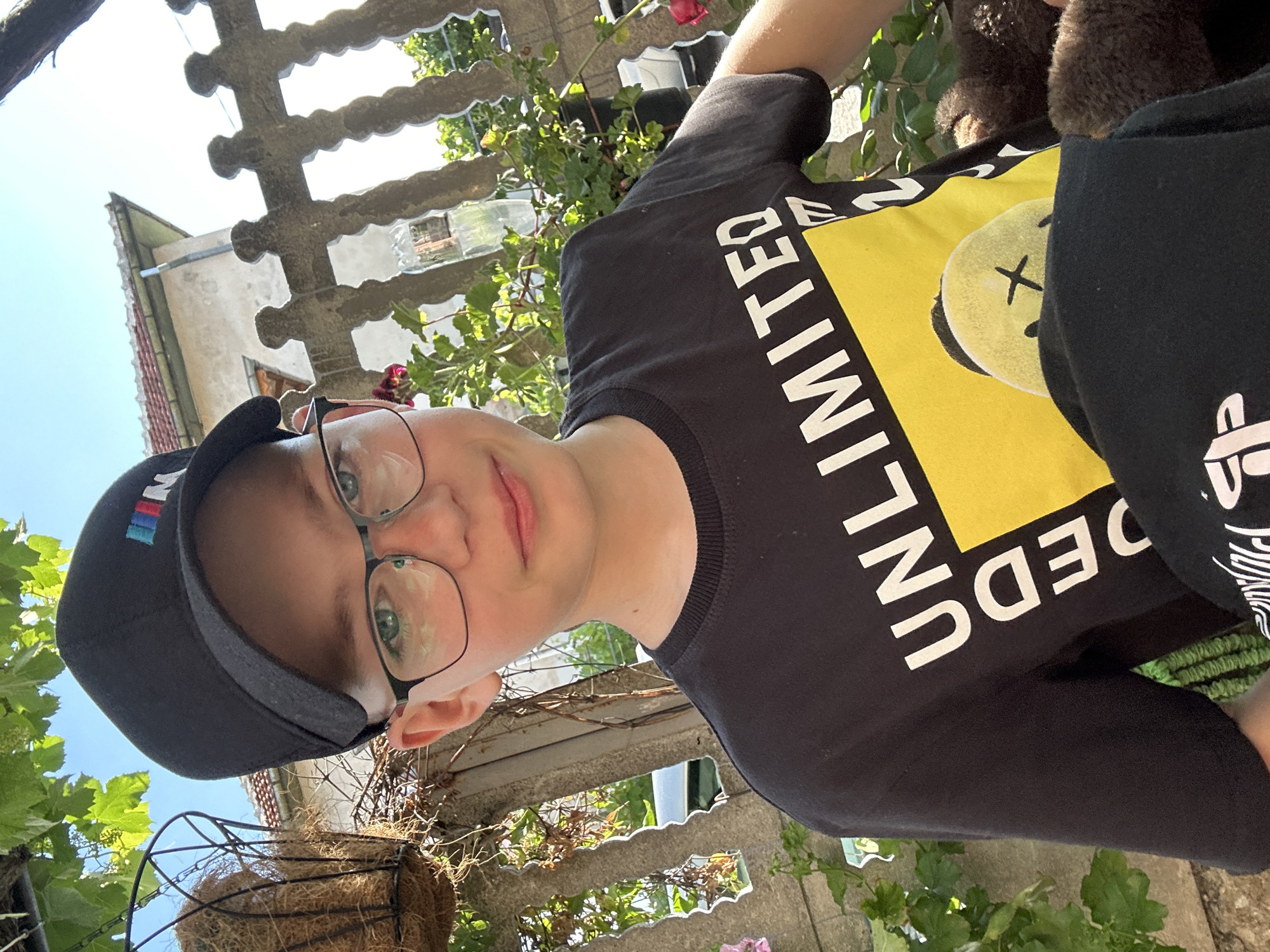 The media file shows a young person wearing a black baseball cap and glasses, taking a selfie outdoors in what appears to be a backyard or garden. The person is wearing a black t-shirt with large white text that reads "UNLIMITED". A yellow square is visible on the t-shirt with a cartoonish face inside. The setting includes a concrete fence or wall with greenery and foliage growing around it. In the background, a building with a red-tiled roof can be seen. The sky is bright blue, suggesting it is daytime with clear weather. The person's facial expression is neutral, and the overall mood seems relaxed. The image is oriented sideways. The visible text on the cap is "BMW" in colorful stripes. The text on the t-shirt is "UNLIMITED".