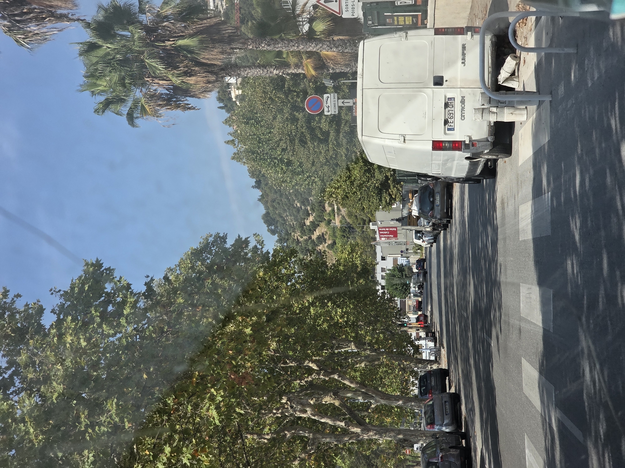 The image shows a sunlit street in Nice, France, with a clear blue sky overhead and lush green trees lining the left side of the road. Several cars are parked along the sidewalk, and a white van is positioned in the right lane, facing away from the camera. The van has the text "CITROEN JUMPER" visible on its rear. A pedestrian crossing is marked on the asphalt in the foreground. In the background, buildings are visible on a hillside, with more trees and vegetation. Traffic signs are present, including a circular red and blue sign indicating parking restrictions and a triangular yellow sign with a red border indicating a warning. The overall scene depicts a typical urban streetscape on a bright day.