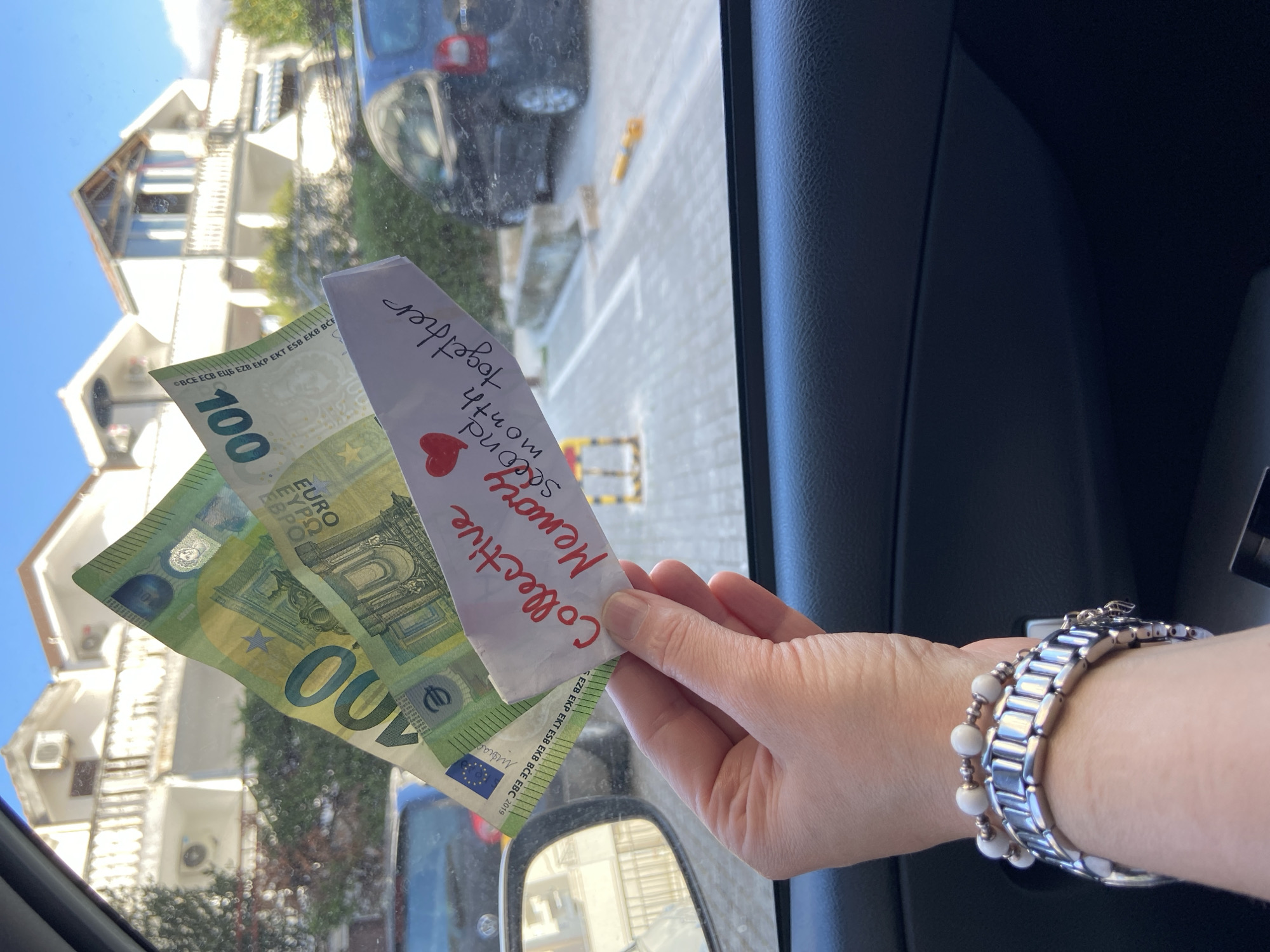 The photograph, likely taken from within a car, captures a hand holding two 100 Euro bills and a small paper note against the backdrop of a street scene, partially visible through the car's window. The note, handwritten, reads "Collective Memory second month together" with a small red heart. The person's wrist is visible, adorned with a silver watch and a beaded bracelet. The view outside the car window includes parts of a building and a car parked on the street. Based on the style of the building visible through the windshield in the background, it is highly likely that the photograph was taken in the city of Budva, Montenegro.