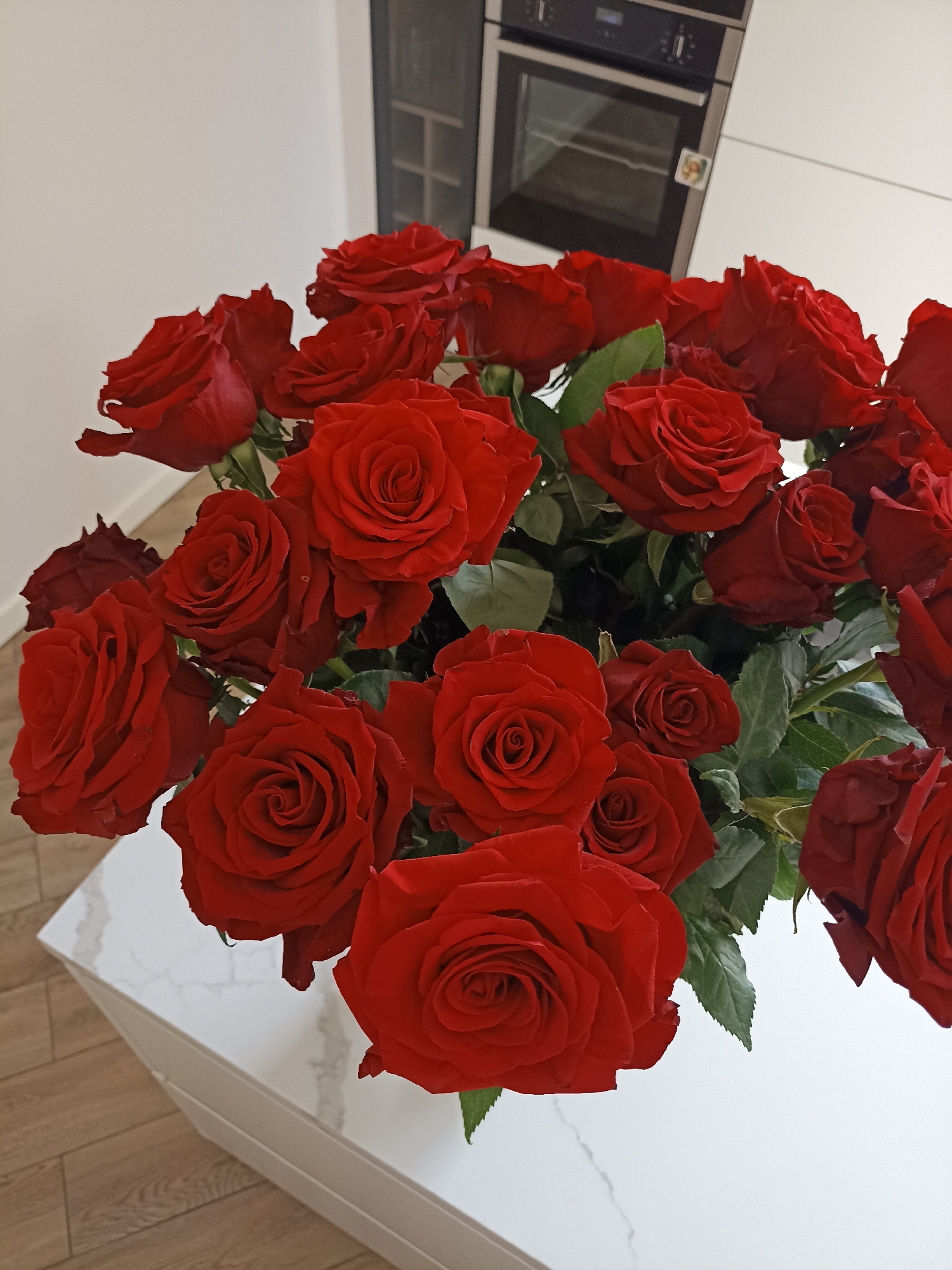 A bouquet of deep red roses is arranged on a white marble-patterned surface, likely a table or countertop, in an indoor setting. The roses are in various stages of bloom, with some fully open and others still tightly furled. Their velvety petals are a rich, saturated red, and they are interspersed with lush green leaves. The background features a portion of a modern kitchen appliance, possibly an oven, and a white cabinet or wall. The lighting suggests an interior environment, potentially a home or a well-lit event space. The overall impression is one of elegance and natural beauty.