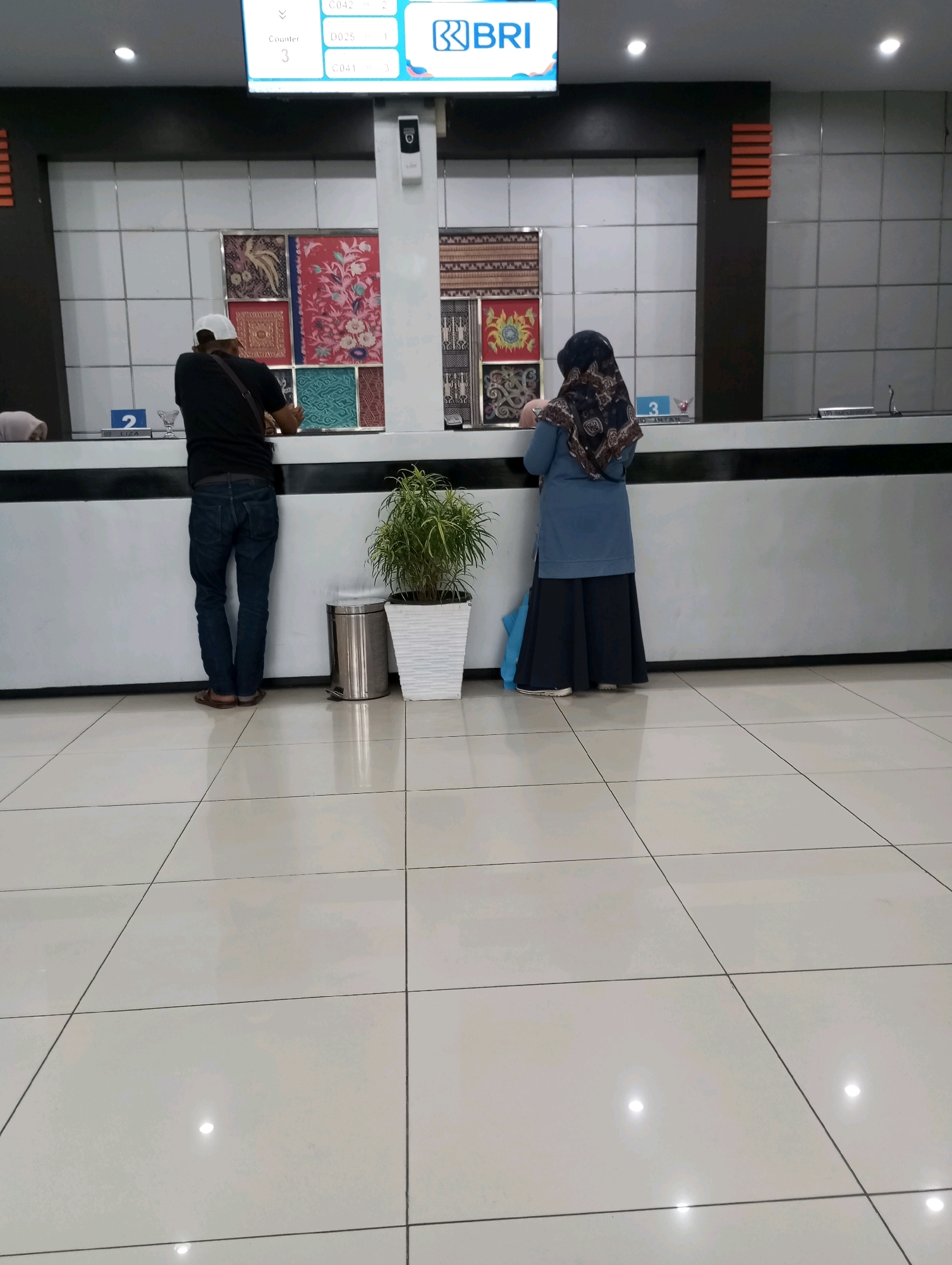 In Padang Panjang, Indonesia, a man wearing a white cap and a black t-shirt stands at a counter in what appears to be a bank or service center. He is looking down at the counter. A woman in a blue hijab and a blue top with a long dark skirt is also at the counter, facing away from the camera. She is interacting with someone behind the counter. Above them, a digital display shows "Counter 3" and "0042", "0025", "0041". A potted plant sits to the right of the man. The environment appears to be indoors with tiled flooring and a counter area. The wall behind the counter is decorated with colorful textile panels.