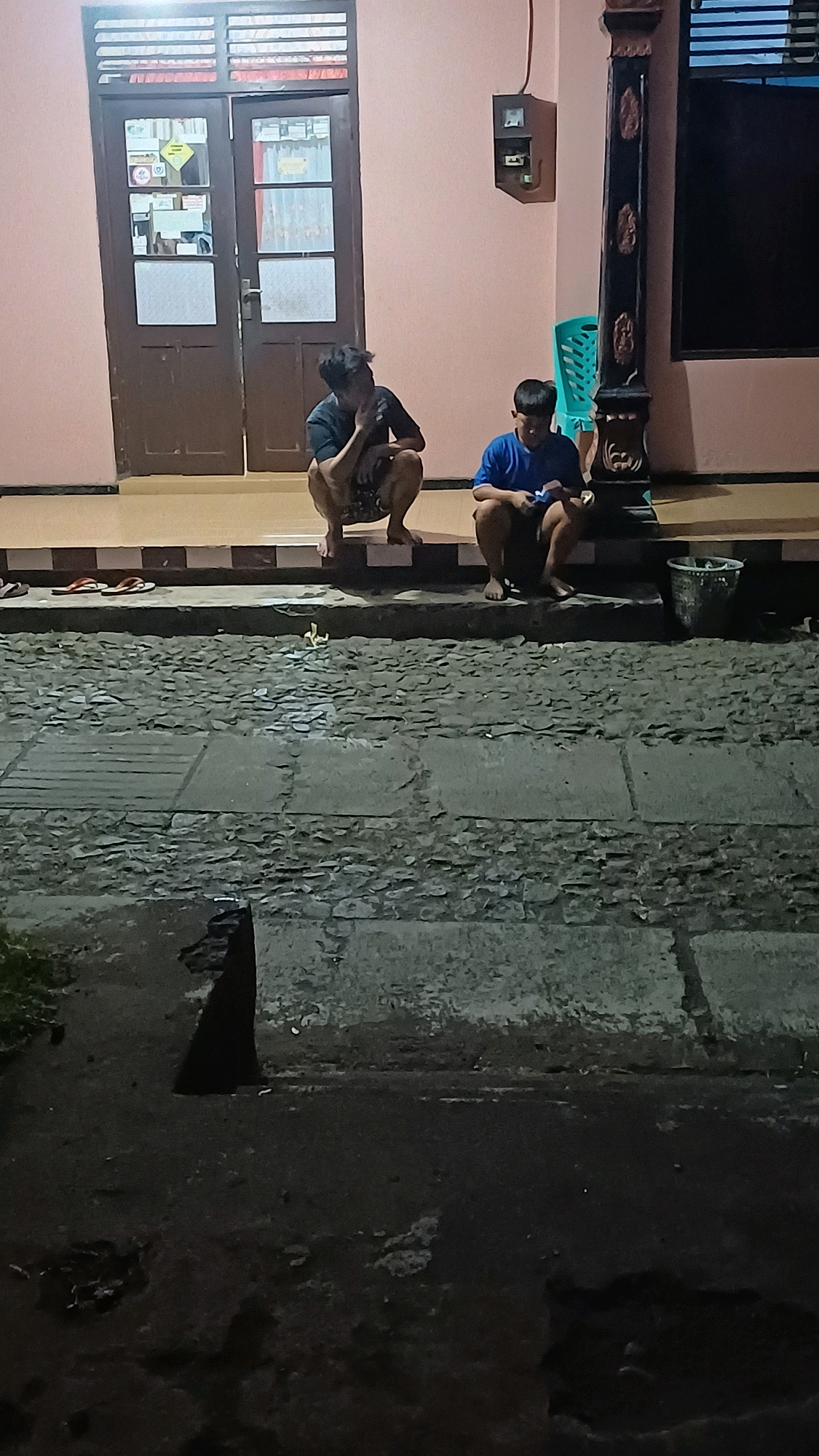 Two young males are squatting on the steps of a building, possibly a residence, in Wonokerso, Indonesia. The male on the left, with dark hair, is wearing a dark t-shirt and patterned shorts and is holding his hand to his face. The male on the right, with shorter dark hair, is wearing a blue t-shirt and dark shorts and is looking down at something in his hands. In front of them is a paved area, which appears to be a walkway or driveway. To the right of the steps, a trash can is visible. The building has double doors with glass panes and a light-colored facade. The scene is dimly lit, suggesting it is nighttime or early morning.