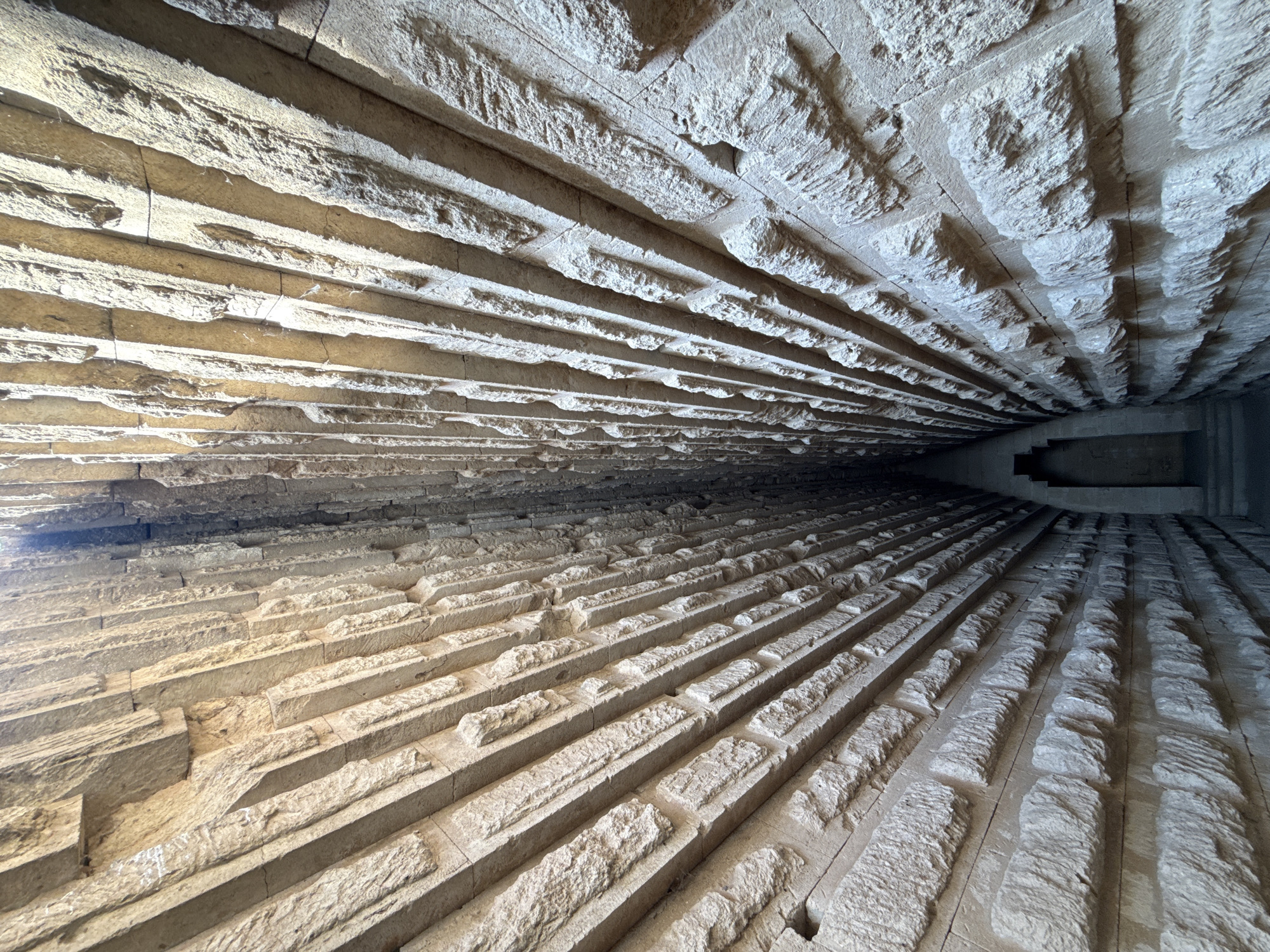 The image captures a view looking down a narrow, elongated passage constructed with roughly hewn stone blocks. The walls of the passage are lined with these blocks, which are arranged in a pattern that gives the impression of converging towards a dark opening at the far end. The stone exhibits a rough texture and varies in color from light beige to a warmer tan, with shadows defining the contours of the individual blocks. Sunlight streams in from the top, illuminating parts of the passage and creating strong contrasts. The passage appears to be an architectural structure, possibly an ancient tomb or tunnel. Given the context of Kerch, Ukraine, this likely depicts a section of the Mithridates Tomb or a related ancient site in the vicinity. There are no people or living beings visible in the image.