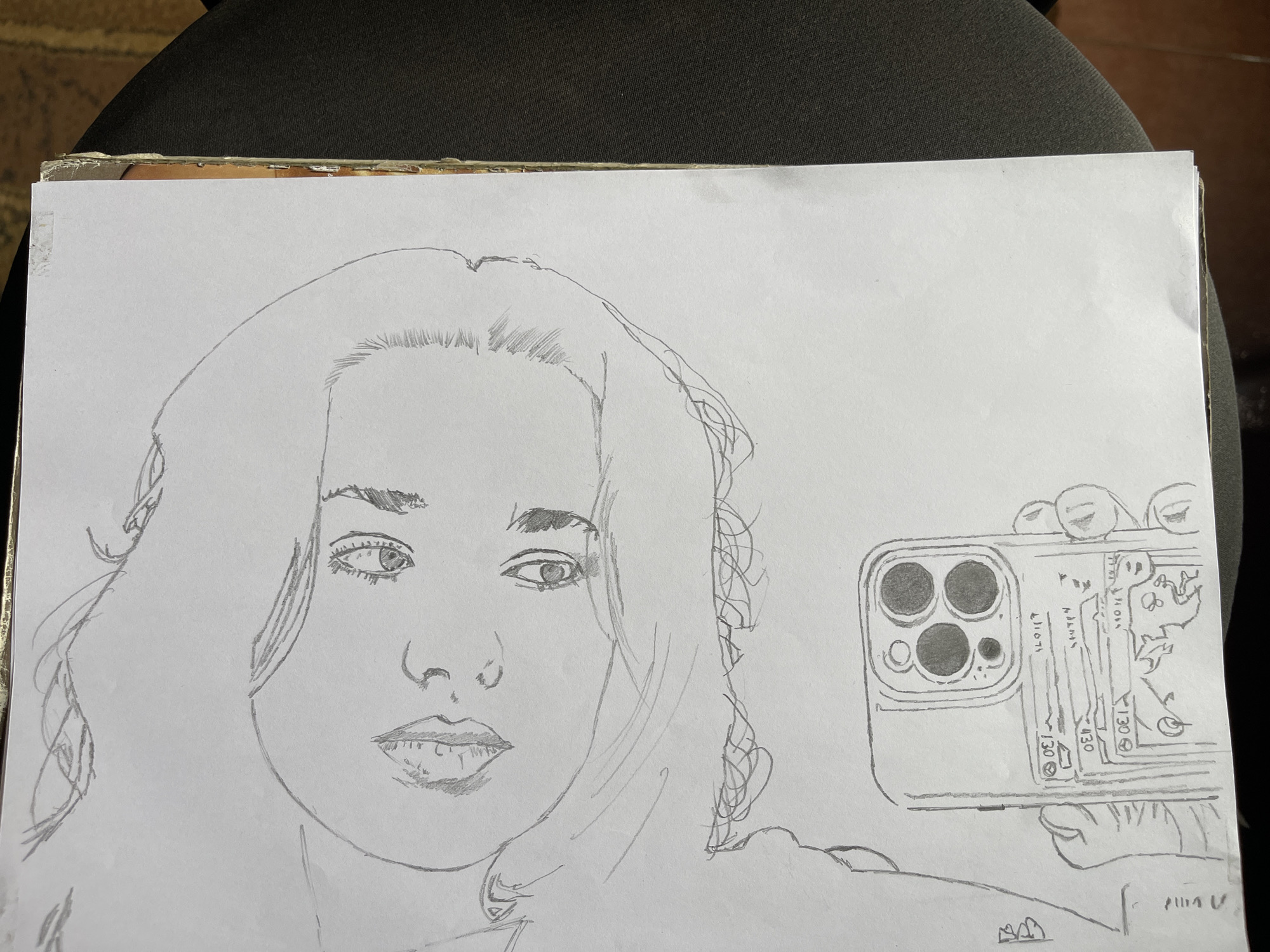 The image shows a pencil drawing of a person taking a selfie with a phone. The subject is a person with shoulder-length hair. The phone has multiple camera lenses and writing and drawings on the back. The drawing is on a piece of paper that is placed on a dark surface. There is no information in the image to determine the location as Jos, Nigeria.