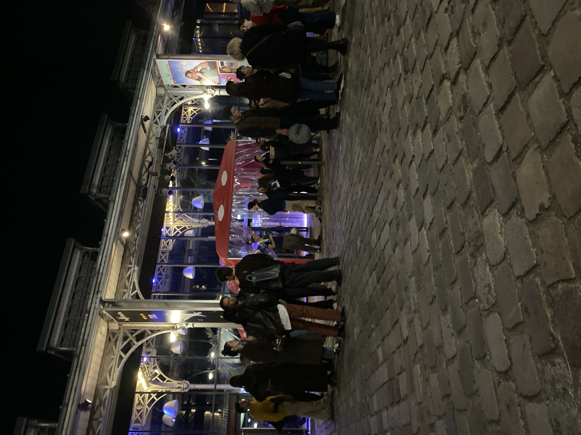 The image depicts a nighttime scene on a cobblestone street in Paris, France. A line of people is waiting outside a building with ornate, Eiffel Tower-like architectural details. The building has large glass windows, exposing interior lighting and decorations. A red and white polka-dotted mushroom-shaped object is visible near the entrance. The people are dressed in casual attire and appear to be waiting in line, possibly for an event or entrance to the building.
