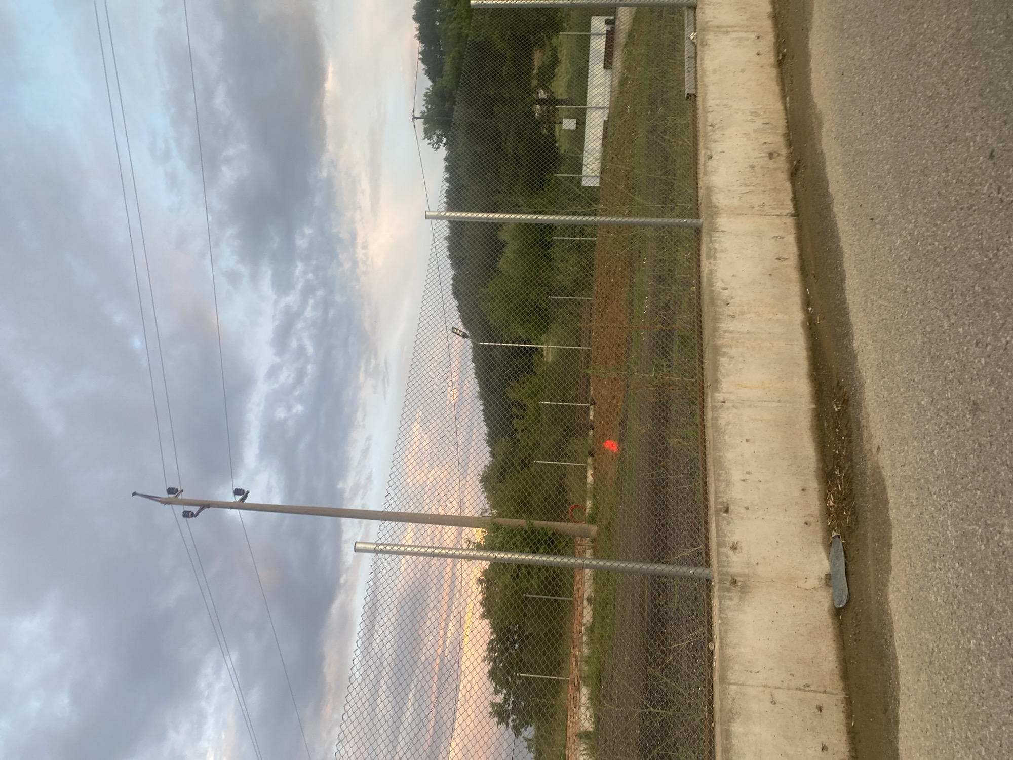 The image depicts an outdoor scene near Ablanitsa, Bulgaria, characterized by a cloudy sky with diffused sunlight. In the foreground, a rough asphalt surface leads to a concrete retaining wall. Beyond the wall, a chain-link fence encloses an area with dry, brown soil and sparse vegetation. A tall, thin electrical pole with insulators and wires extends vertically on the left side of the frame, passing through the fence. Further back, dense green foliage forms a hillside under the overcast sky. A small, bright orange cone is visible on the ground within the fenced area, and a white rectangular structure is present in the middle distance to the right. The overall impression is one of a construction or undeveloped area on the outskirts of a town.