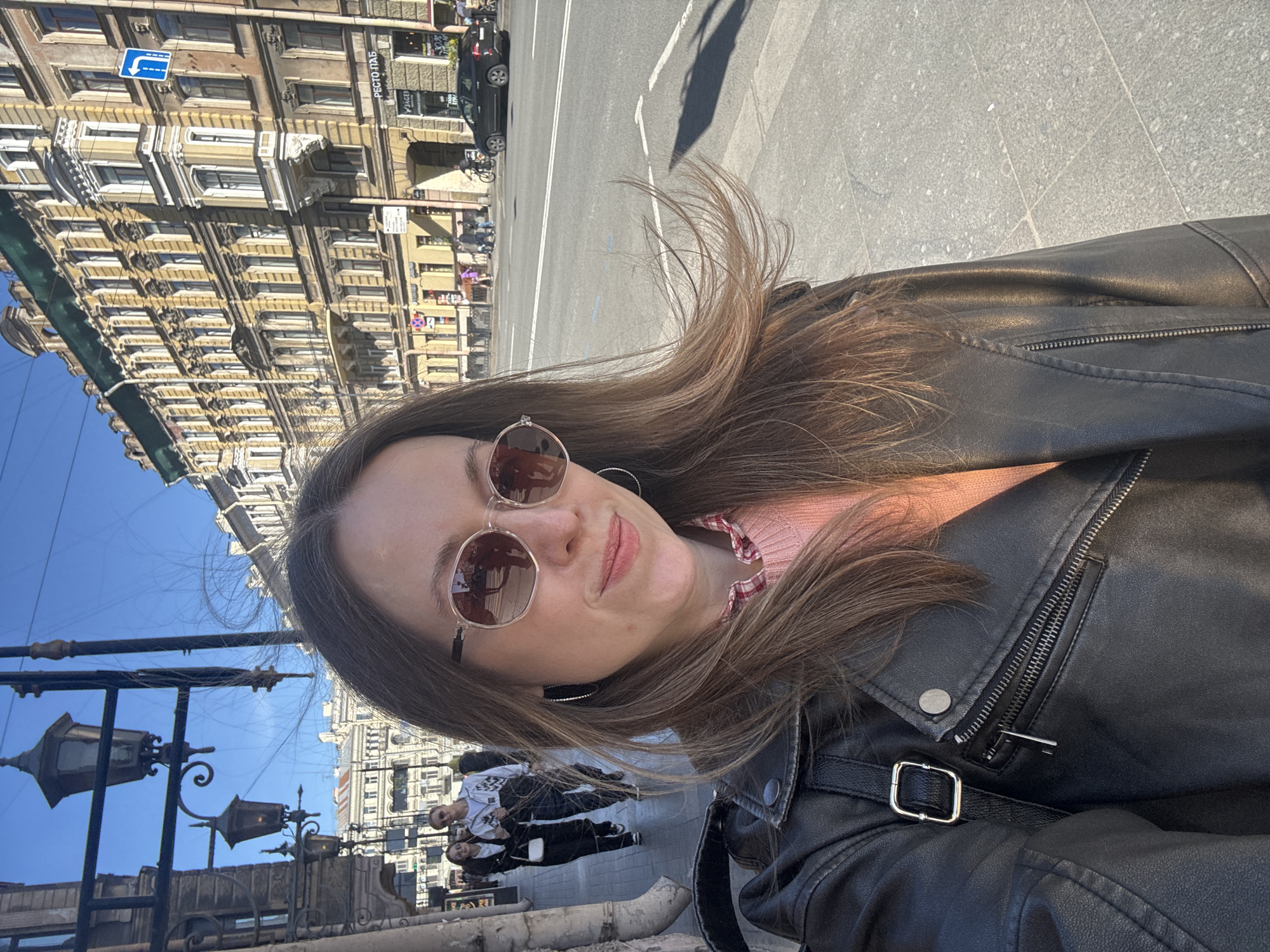 A woman wearing sunglasses and a black leather jacket stands outdoors in Saint Petersburg, Russia. She is likely taking a selfie. In the background, a street with a multi-story building featuring ornate architectural details is visible. A street lamp is present in the lower left of the frame. A car drives down the street in the background. Other people are seen walking on the sidewalk. The weather appears sunny.