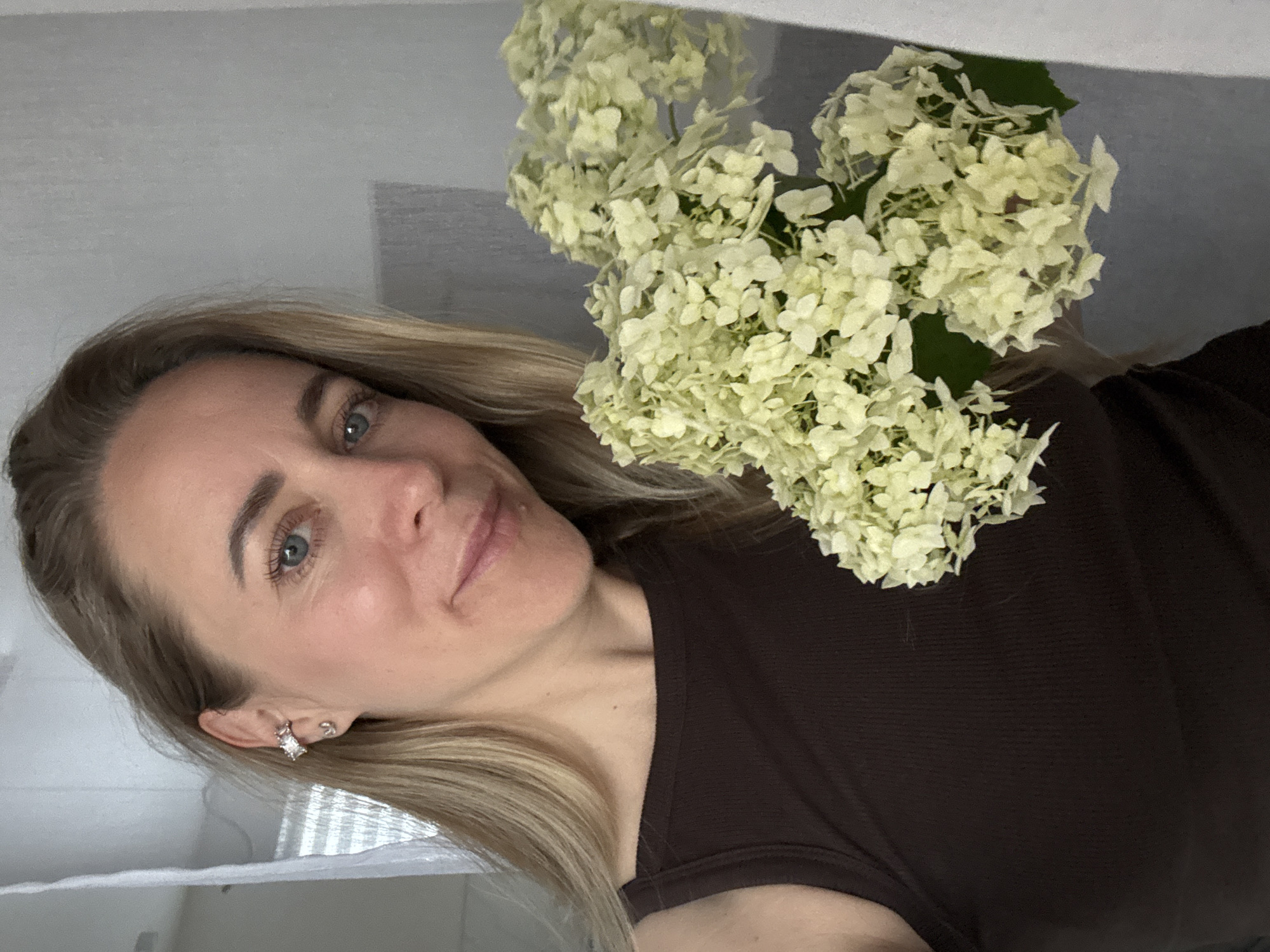 In a close-up, indoor shot, a woman with blonde hair and blue eyes smiles at the camera, holding a bouquet of light-colored hydrangeas. She is wearing a dark brown tank top and a sparkly earring in her left ear. The background appears to be a neutral-colored wall. The image does not provide any contextual information that would allow for a determination of its location within the city of Chaikovsky, Russia.
