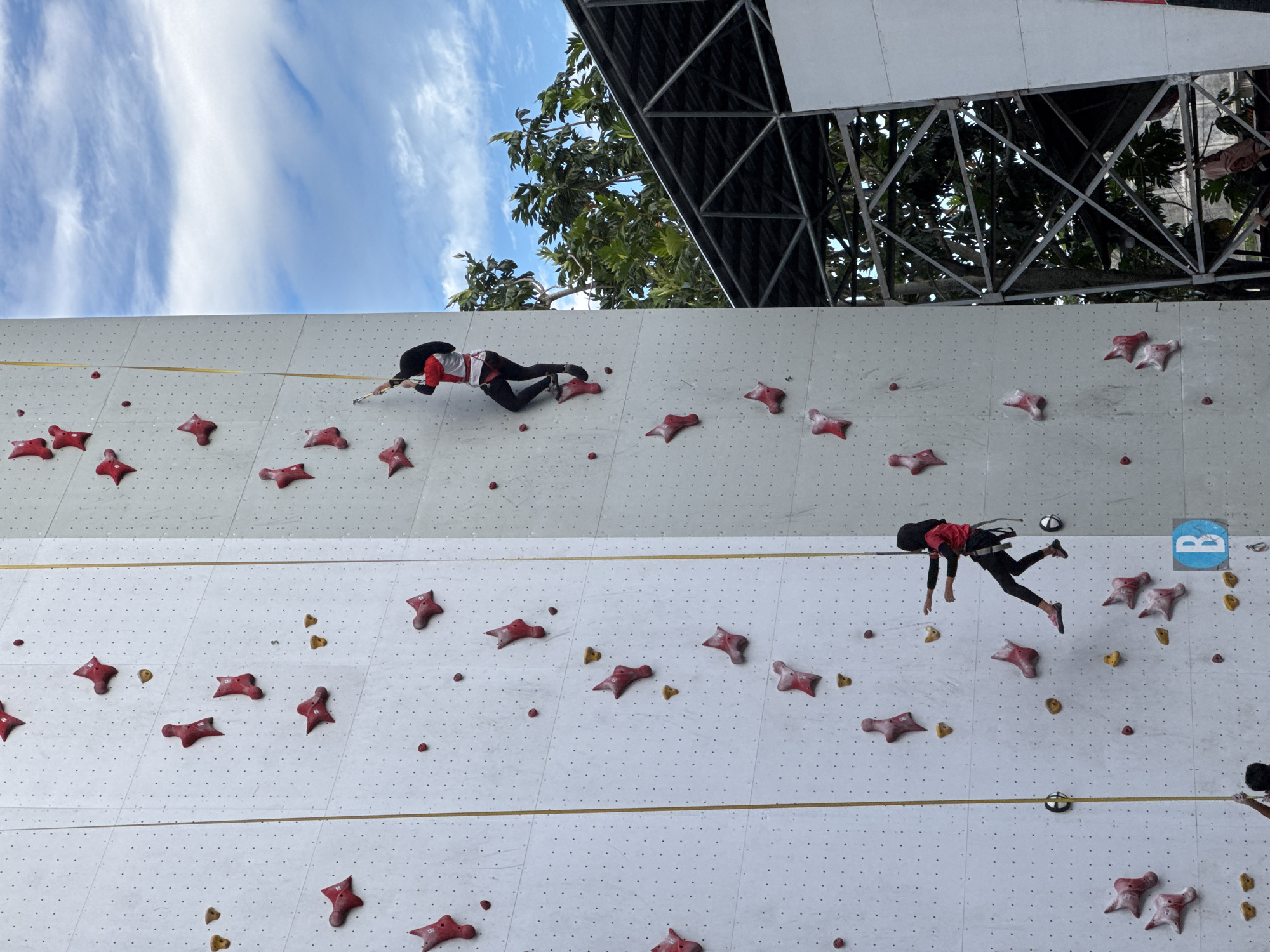 In Banda Aceh, Indonesia, two individuals are rock climbing on an outdoor climbing wall. The wall is white with numerous red and yellow climbing holds. The climber on the right, wearing a red and white shirt and black leggings, is higher up on the wall. The climber on the left, dressed in a similar red shirt and black leggings, is positioned lower on the wall. Both climbers are secured by ropes. The climbing wall is situated outdoors, with a blue sky and scattered clouds visible above. Lush green trees and a metal structure are present in the background, suggesting an athletic facility or park.