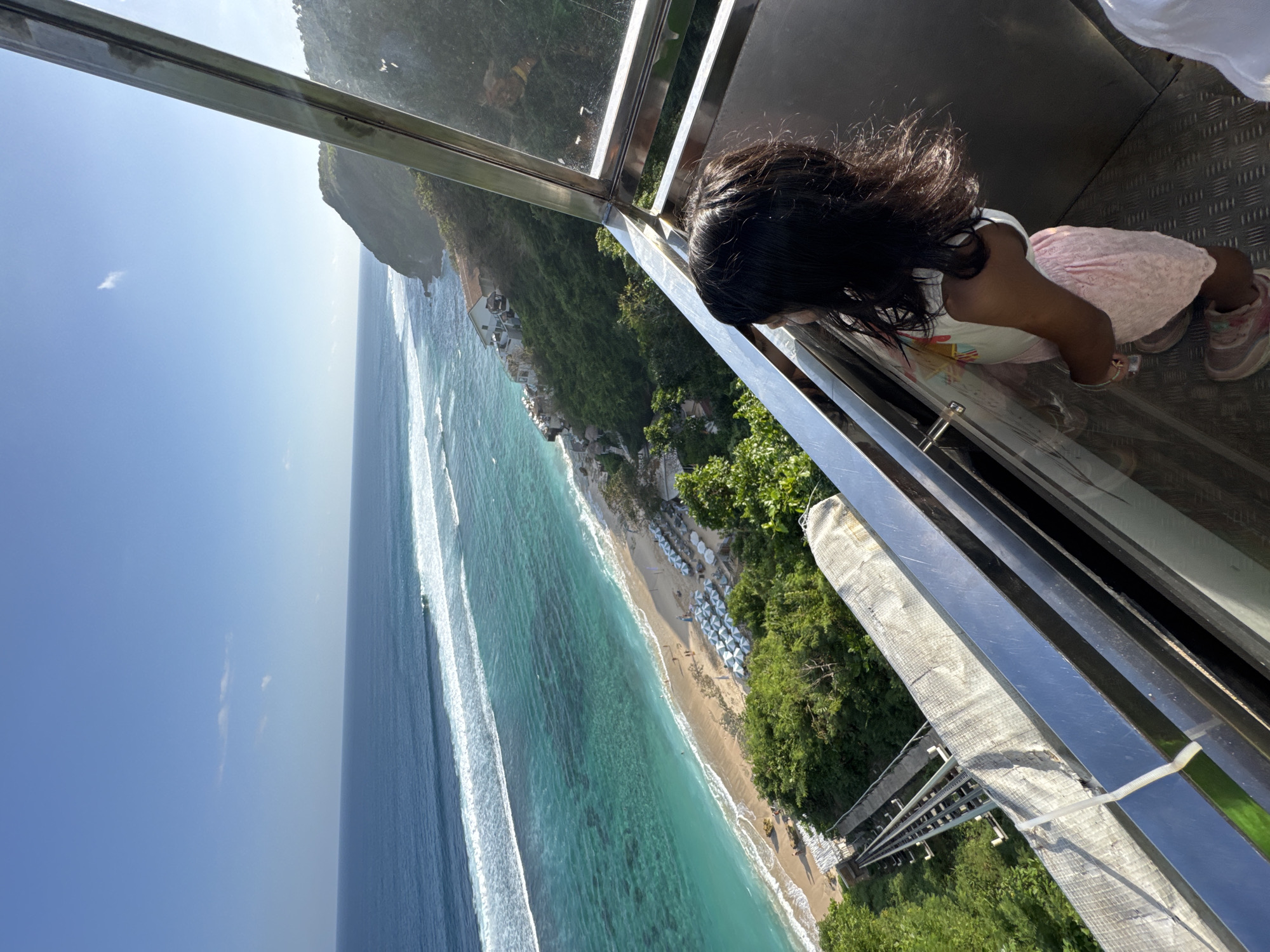 In Kuta Selatan, Indonesia, a young girl stands inside a glass and metal elevator, looking out at a scenic coastal view. The elevator car has a checkered metal floor and polished metal railings. The girl, with dark hair, wears a white top with a colorful print and a light pink skirt. She is looking towards the ocean, which is a striking turquoise color near the shore and deep blue further out. White waves crash onto a sandy beach lined with rows of light blue beach umbrellas and lounge chairs. Lush green vegetation covers the hillside leading down to the beach. A funicular railway track is visible on the left side of the image, descending the slope. The sky above is a clear, bright blue with a few wispy clouds. The overall scene captures a beautiful tropical beach landscape viewed from an elevated perspective.