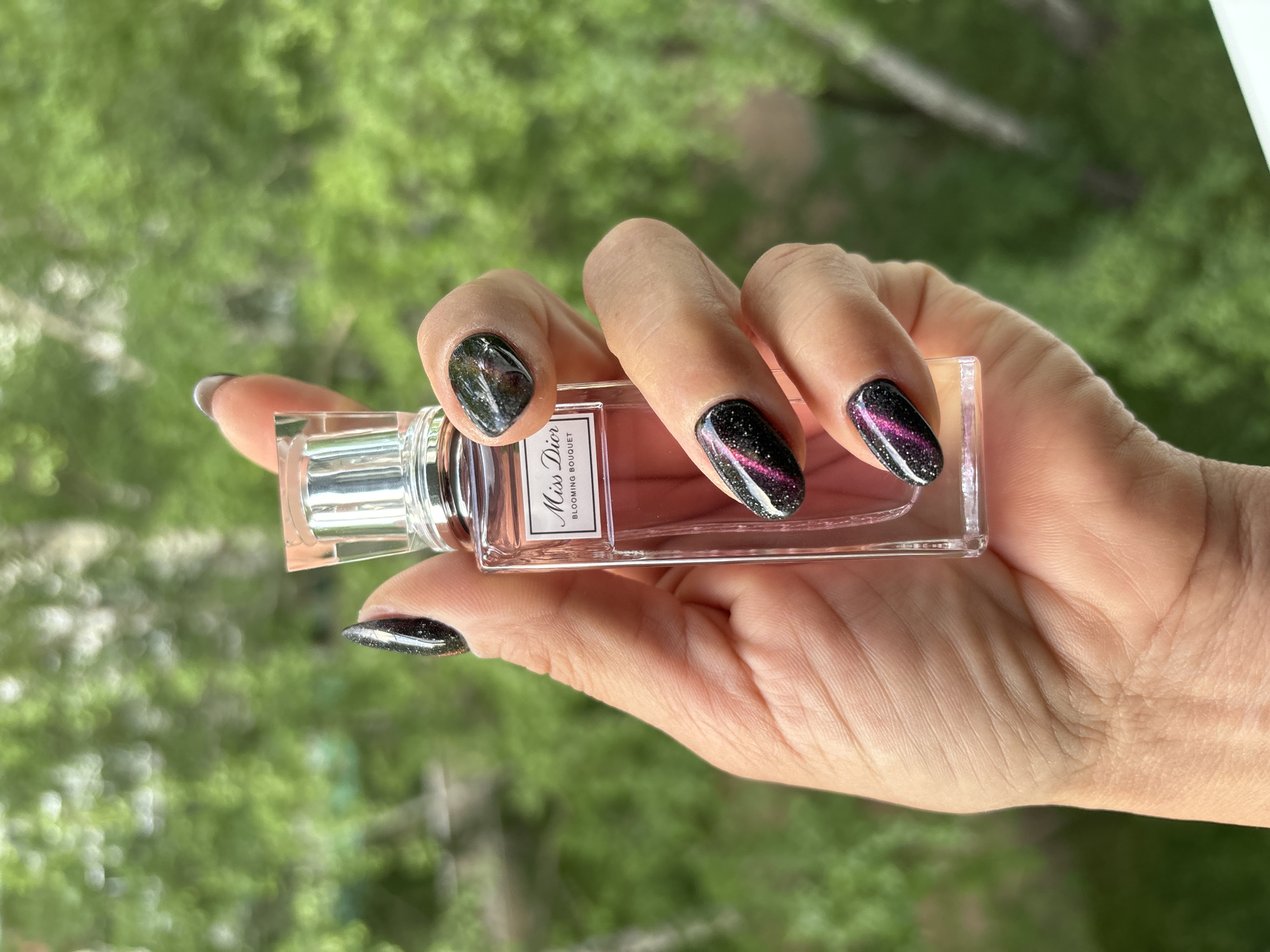 A close-up, vertical, outdoor shot shows a hand holding a small, rectangular bottle of Miss Dior Blooming Bouquet perfume. The bottle is made of clear glass and has a silver cap. The perfume inside is a light pink color. The hand has manicured nails painted with a dark, glittery polish that has purple and pink streaks. The background is blurred, showing green foliage, suggesting the photo was taken outside with trees or plants. The lighting is natural daylight. There are no discernible landmarks indicating a specific location within Chaikovsky, Russia.