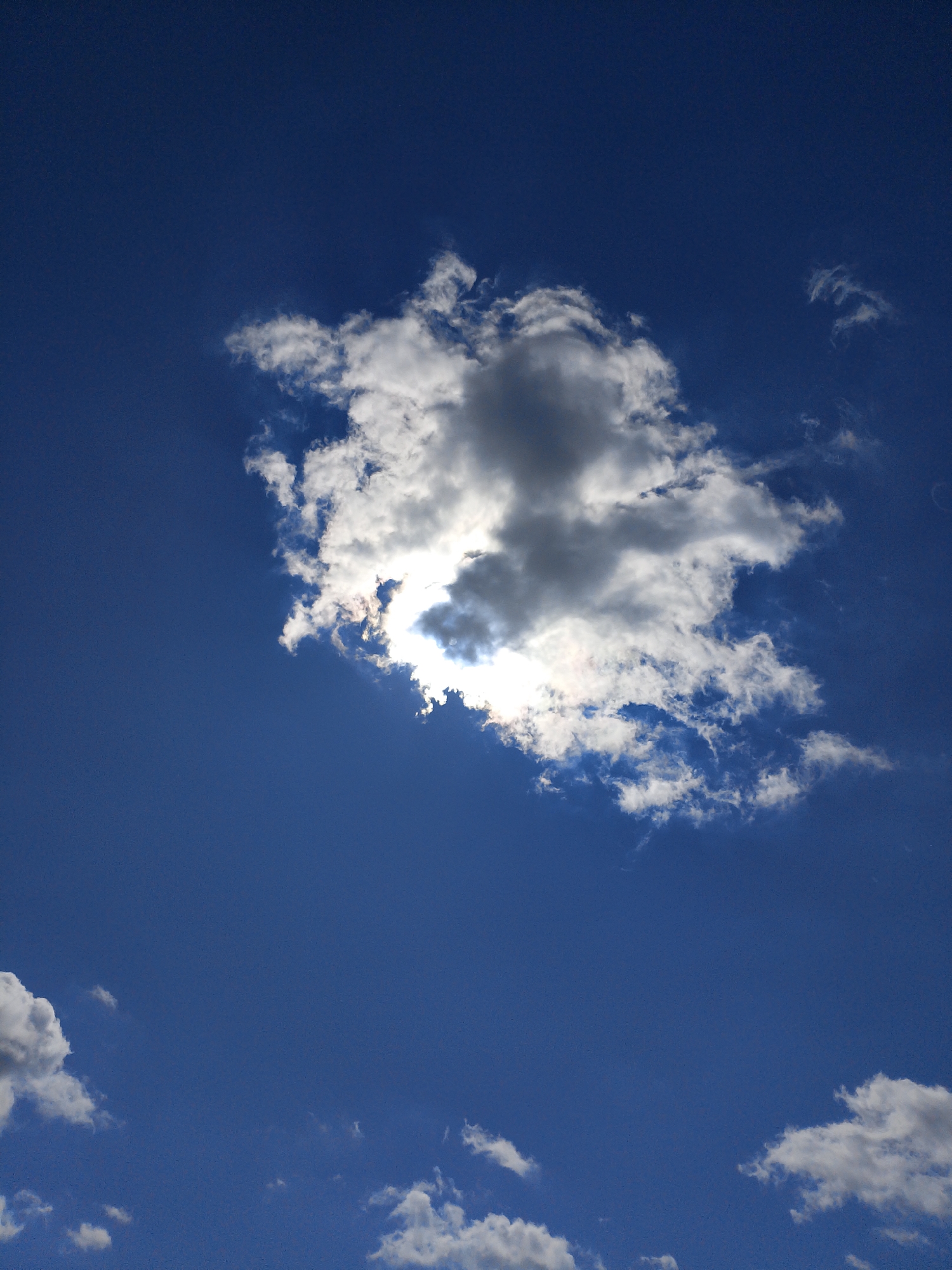A bright, sunlit cloud is partially obscuring the sun against a deep blue sky. The cloud is dense with both bright white and darker gray portions, indicating varying thickness and light penetration. Smaller, wispy clouds are scattered across the sky. The image is a close-up shot, focusing entirely on the sky and clouds, with no discernible city landmarks or environmental context. The scene appears to be outdoors during daytime.
