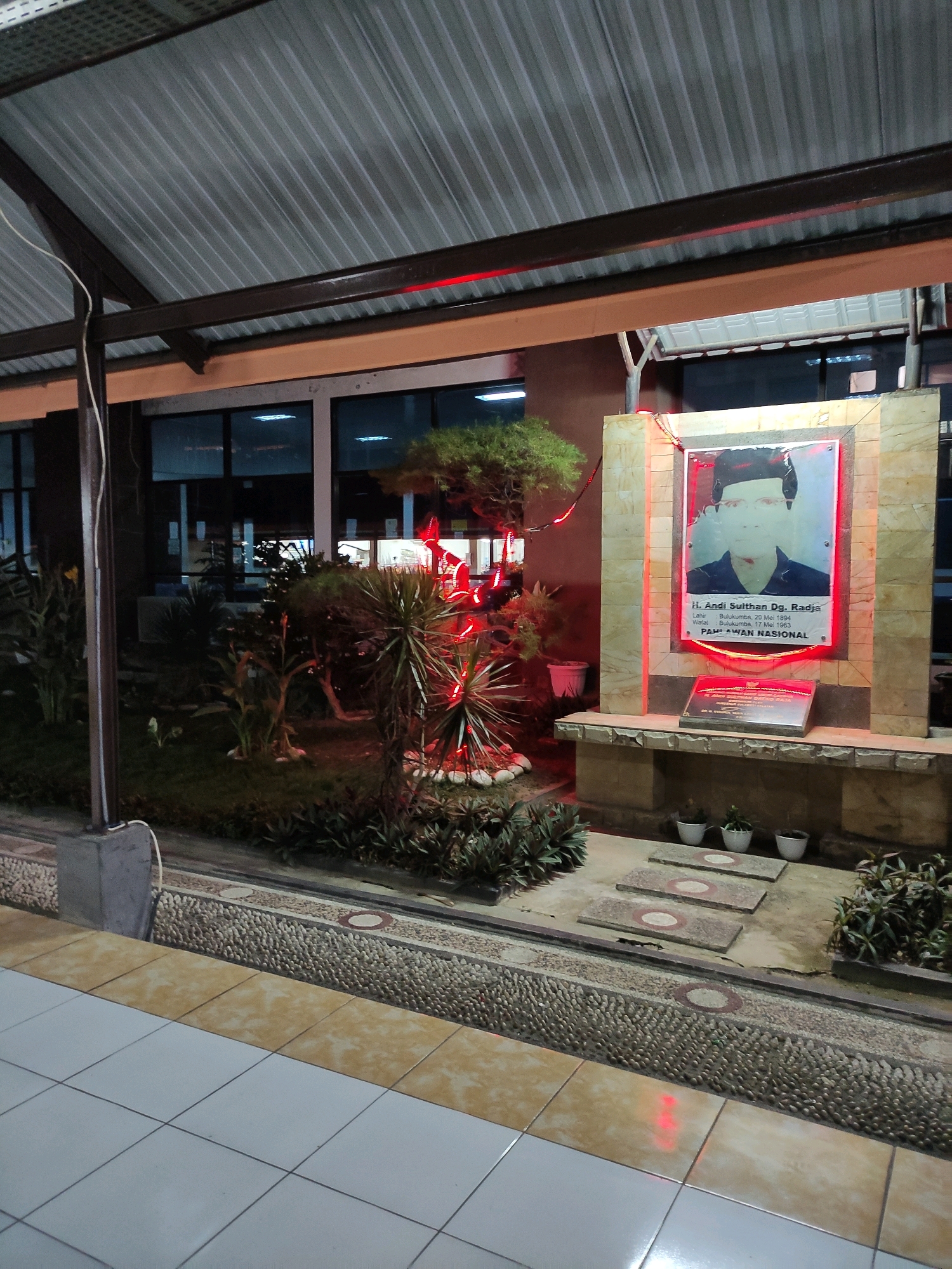 The image depicts a memorial monument at night, located outdoors. The monument features a framed portrait of a man identified as H. Andi Sulthan Dg. Radja, a "Pahlawan Nasional" (National Hero). Text below the portrait provides his birth and death dates: "Lahir: Bulukumba, 20 Mei 1894, Wafat: Bulukumba, 17 Mei 1963." The monument is adorned with red string lights. In front of the monument, there is a smaller plaque with text that is not fully legible. The monument is situated in a landscaped area with various plants, including a small tree and what appear to be spiky plants. There are also stepping stones leading towards the monument and several small potted plants. The background shows a building with large windows, and the scene is illuminated by artificial lighting, including red and white lights, and the ambient light from within the building. The foreground reveals a tiled walkway. The presence of the name "Bulukumba" on the monument indicates that this memorial is located within or related to the city of Bulukumba, Indonesia.