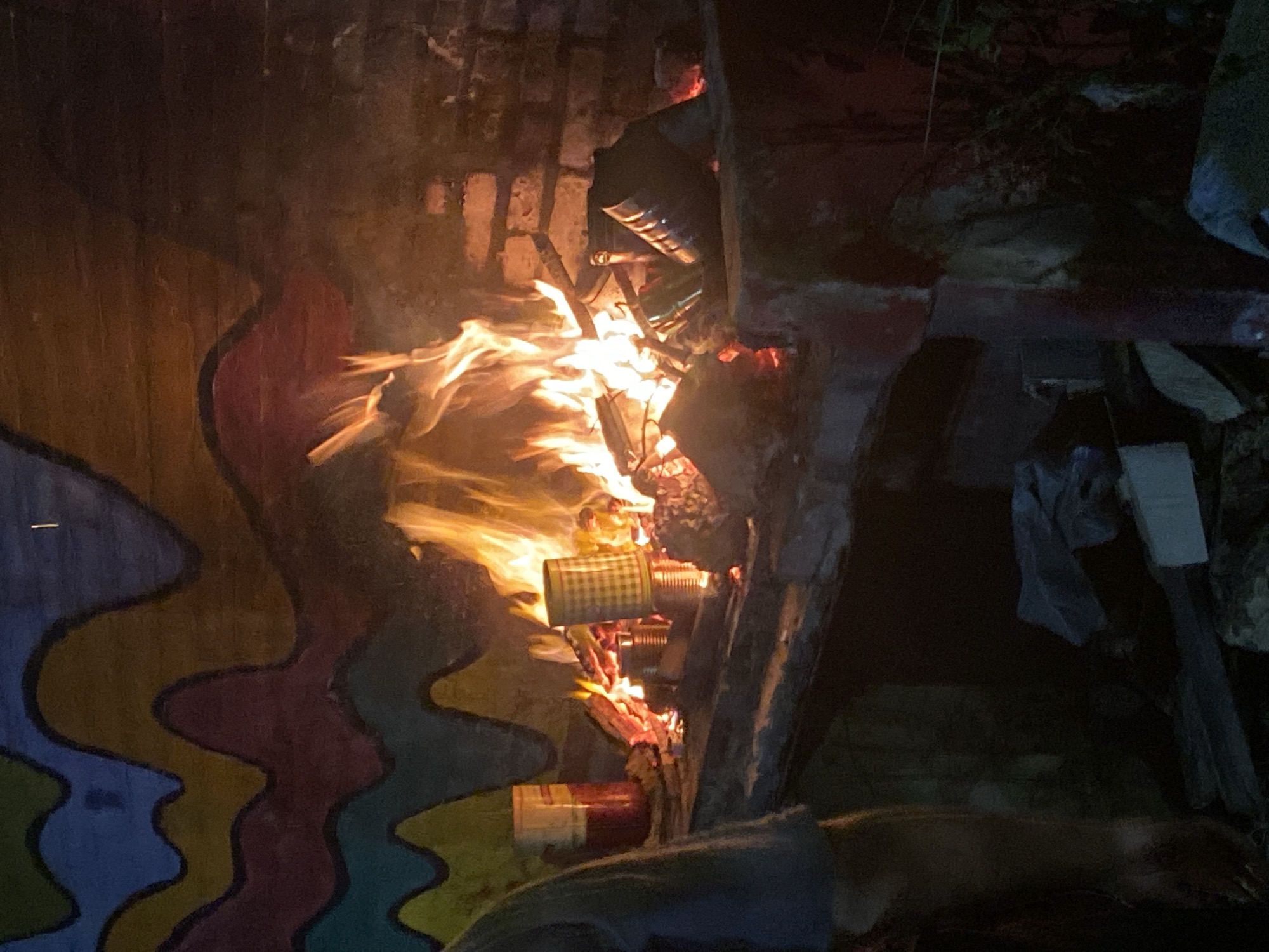 The image depicts an intense fire burning at night, captured in a portrait orientation where the flames spread vibrantly towards the upper right. The fire is characterized by bright orange and yellow hues, illuminating its immediate surroundings. Within the heart of the blaze, several objects are being consumed: notably, various metal cans are visible, some exhibiting a checkered pattern (e.g., beige and white squares), while others are plain metallic or red. These cans, along with pieces of wood or other debris, appear to be acting as fuel.
In the lower-left section of the image, a vertical surface, likely a wall or partition, is decorated with a colorful, abstract mural. This mural features distinct wavy lines in shades of blue, yellow, red, and green, adding a contrasting artistic element to the scene. The upper-right portion of the image reveals darker, less defined structures, possibly composed of brick, concrete, or assorted debris and discarded materials like fabric or plastic, receding into the shadows.
The setting is distinctly nocturnal, with the fire serving as the sole significant light source against a dark background. No individuals are visible within the frame. The overall impression is of an outdoor fire, potentially a bonfire or a utilitarian fire, set within an informal or urban outdoor environment. No discernible text is visible on any of the objects present. The context of Buenos Aires, Argentina, suggests an urban or semi-urban location for this scene.
