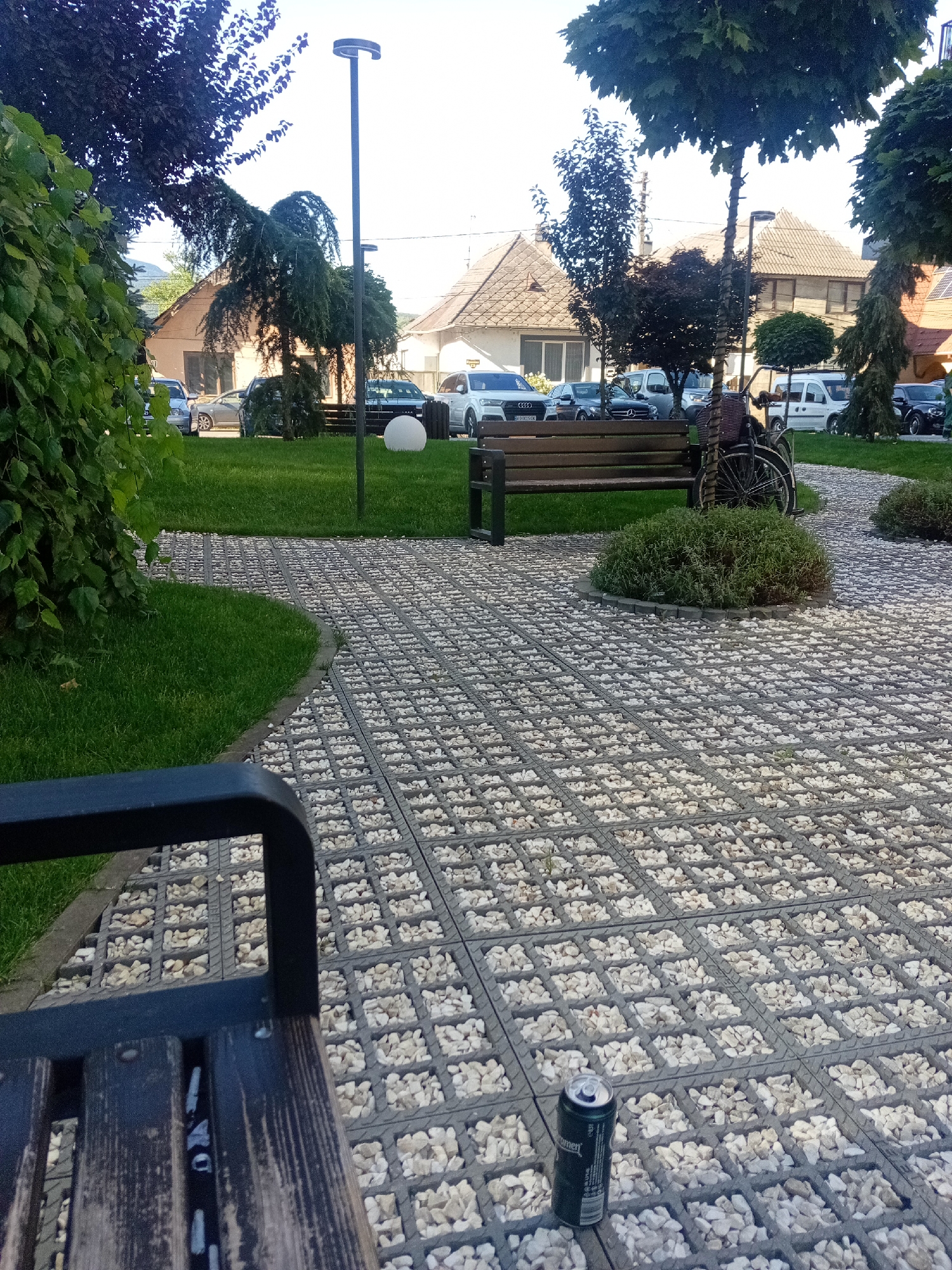 The image depicts a paved area with a gravel infill grid pattern, likely a pedestrian walkway or plaza, in Tiachiv, Ukraine. In the foreground, the armrest and wooden slats of a dark bench are visible. To the right of the bench, a dark beverage can rests on the textured pavement. Further back, a wooden park bench is situated in front of a well-maintained lawn. To the right of this bench, a bicycle with a basket is leaning against a tree. The background features houses with tiled roofs and several parked cars. Trees with lush green foliage are interspersed throughout the scene, and a lamppost stands prominently near the center. The overall impression is that of an outdoor public space, possibly a park or a communal area within a residential district.