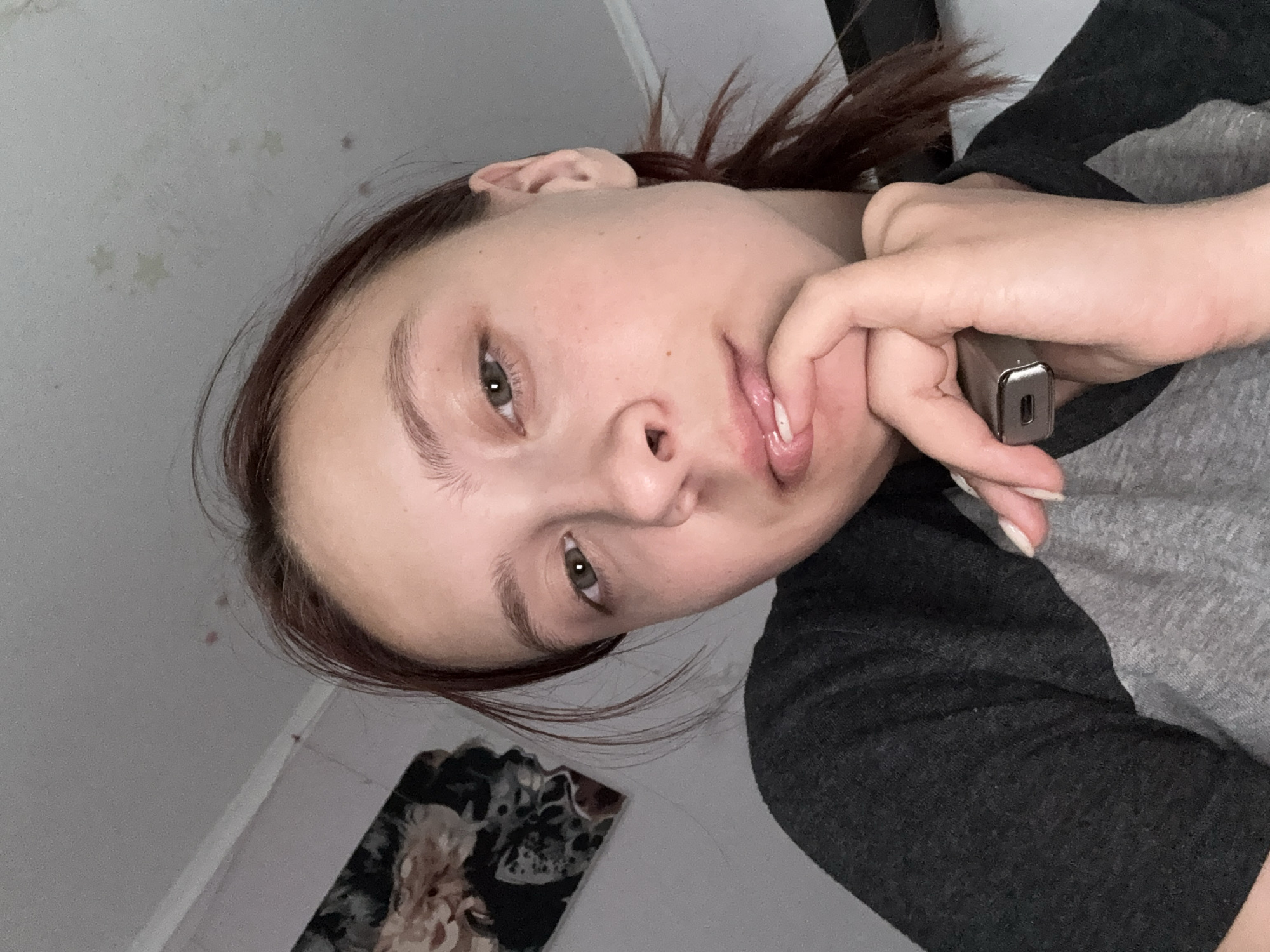 A young woman with brown hair pulled back in a ponytail, wearing a grey and dark grey raglan shirt, is looking directly at the camera. Her right hand is supporting her chin, with her fingers touching her lower lip. She is holding a silver electronic device in her left hand. The background consists of a light-colored ceiling with some dark marks and a wall with a framed picture of flowers. The setting appears to be an indoor, domestic environment, likely a bedroom or living area, within the city of Prikamsky, Russia.