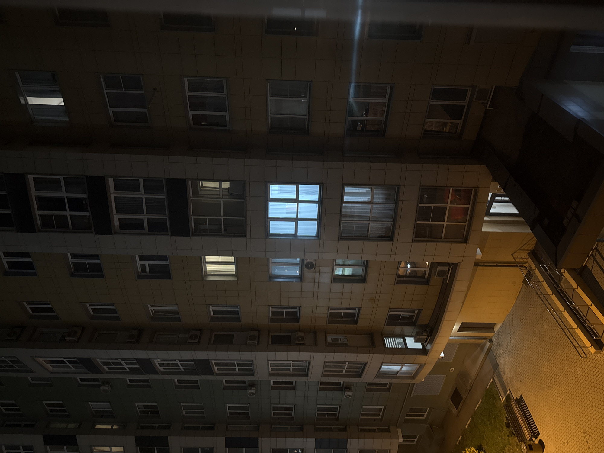 The image depicts an upward view of a multi-story building at night. The building appears to be residential, characterized by rows of windows, some illuminated, and air conditioning units affixed to the exterior. The building's facade is predominantly a light beige or gray color. To the right, a narrow space reveals a section of a courtyard with paved ground and minimal vegetation. The photograph, taken in Khimki, Russia, shows a slice of urban life during the evening. No people are visible in the image.