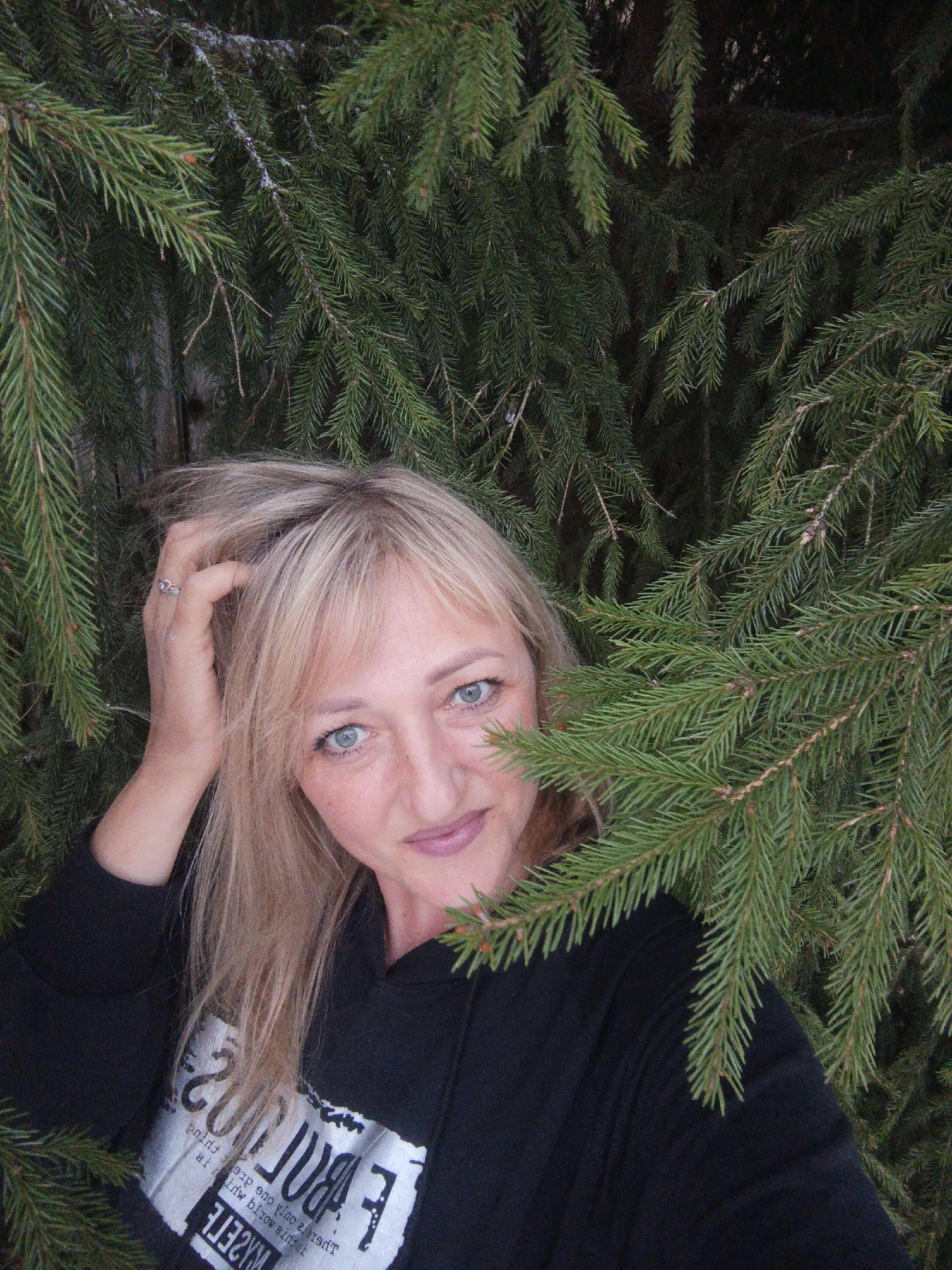 A woman with blond hair and blue eyes is positioned behind evergreen tree branches. She is wearing a black hooded sweatshirt with white text. Her right hand is raised, touching her hair. The location is the city of Chaikovsky, Russia.