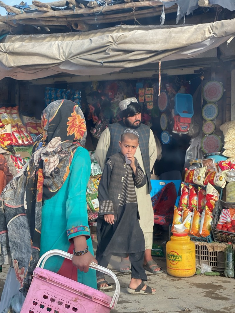 The image shows a street scene in Mir Ahmad Kalay, Afghanistan. A woman wearing a turquoise dress and a headscarf with a floral print stands in the foreground, carrying a pink plastic basket. Next to her, a young boy wearing a dark robe and sandals stands with his finger in his mouth. Behind him, a man with a beard and a traditional cap is partially visible. They are standing in front of a small roadside shop displaying a variety of goods, including snacks, toys, and household items. The shop has a makeshift awning and appears to be located in an open-air market setting.