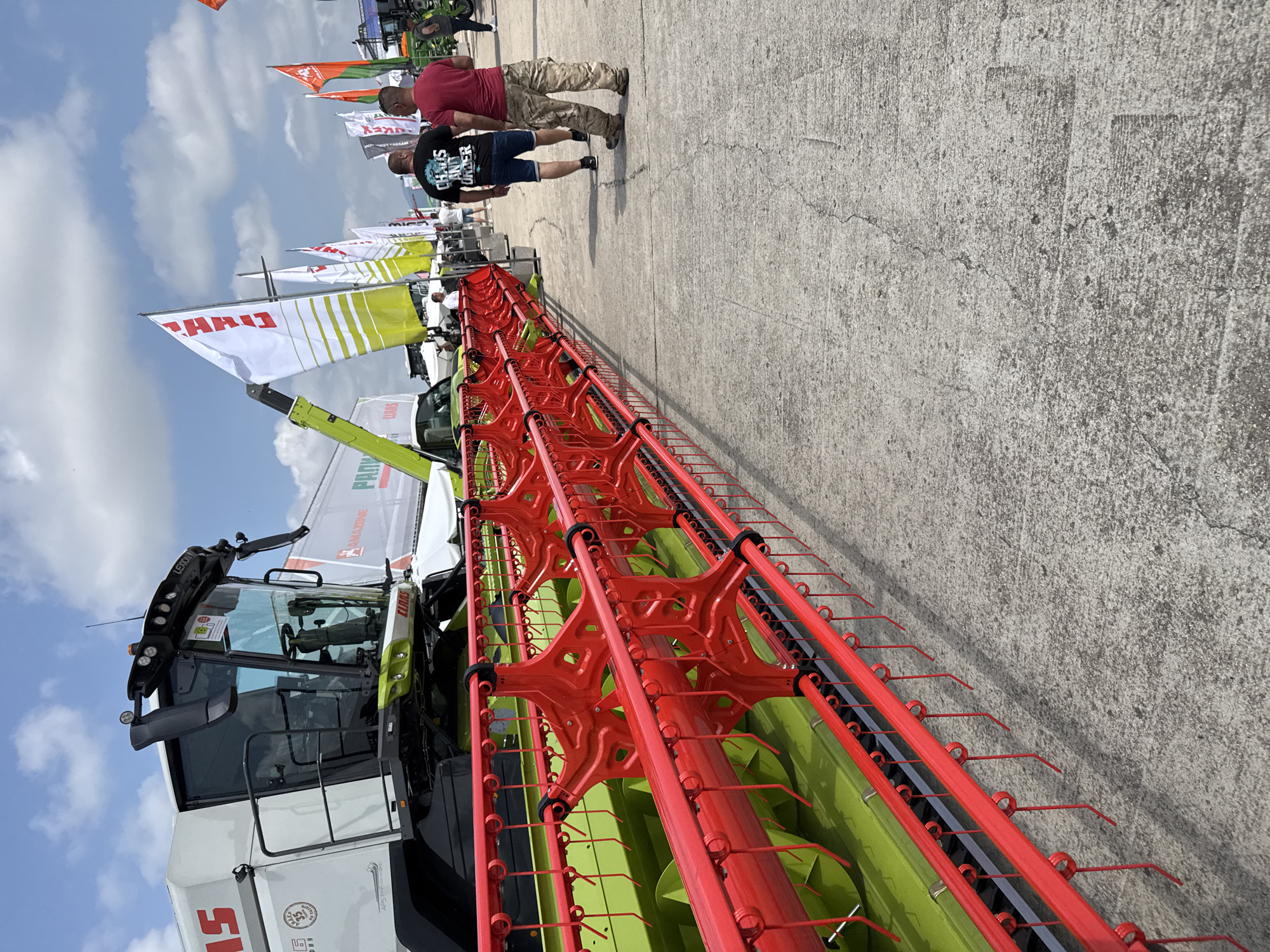 An outdoor scene captured during daytime, likely at an agricultural machinery exhibition in Stara Zagora, Bulgaria. The foreground and left side of the image are dominated by a large, modern CLAAS LEXION combine harvester. Its body is predominantly white, featuring green internal components and a wide, bright red header with numerous tines or fingers. Visible text on the harvester includes "CLAAS", "LEXION", and "APS SYSTEM", along with a circular "50 Years of AXIAL FLOW" logo (partially visible) and some Cyrillic characters.
In the mid-ground, two men walk along a concrete pathway, observing the exhibits. One man wears a red t-shirt and camouflage trousers, while the other wears a black t-shirt with "CHAOS AND ORDER" text and denim shorts. Behind them, several flags, primarily white and green, stand erected. "CLAAS" is clearly visible on multiple flags, with "AMAZONE" and partially "PATRIOT" also discernible.
The scene is set under a bright blue sky with scattered white clouds, indicating clear, sunny weather. The ground is a textured concrete surface with visible cracks. The overall impression is a casual and active outdoor trade show or display of agricultural equipment.