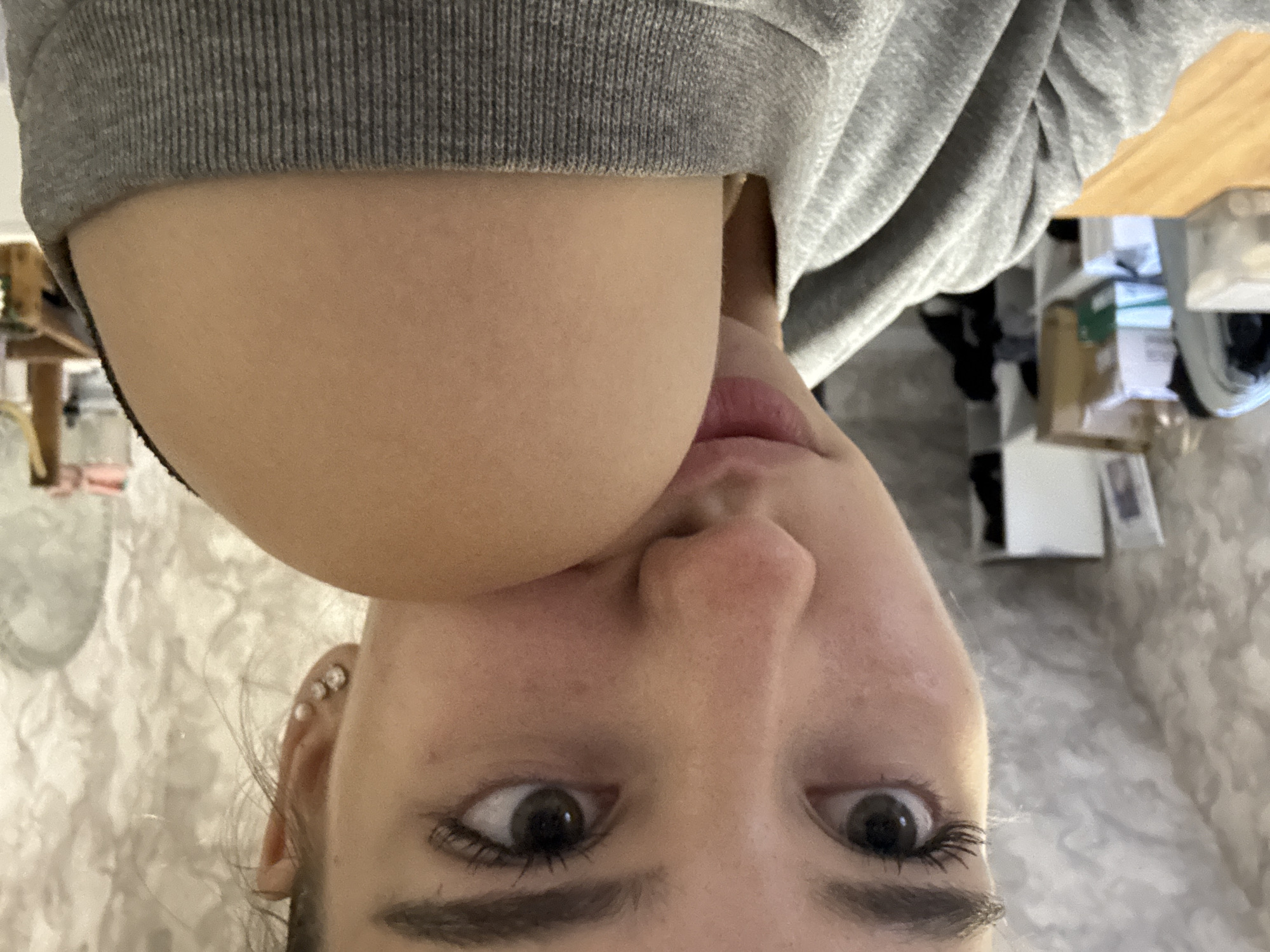 The image is a close-up, selfie-style photograph of a young woman. She is wearing a grey hooded sweatshirt and has her right arm bent, with her shoulder and upper arm resting near her face. Her left shoulder is also visible, partly covered by the sweatshirt. She has dark hair, visible eyelashes, and is wearing small stud earrings in her left ear. Her gaze is directed slightly away from the camera. The background appears to be an indoor setting, possibly a room, with what looks like a textured wallpaper on the wall. There are some objects visible in the blurred background, including shelves with boxes and other items. The location is stated to be Noviy, Russia.