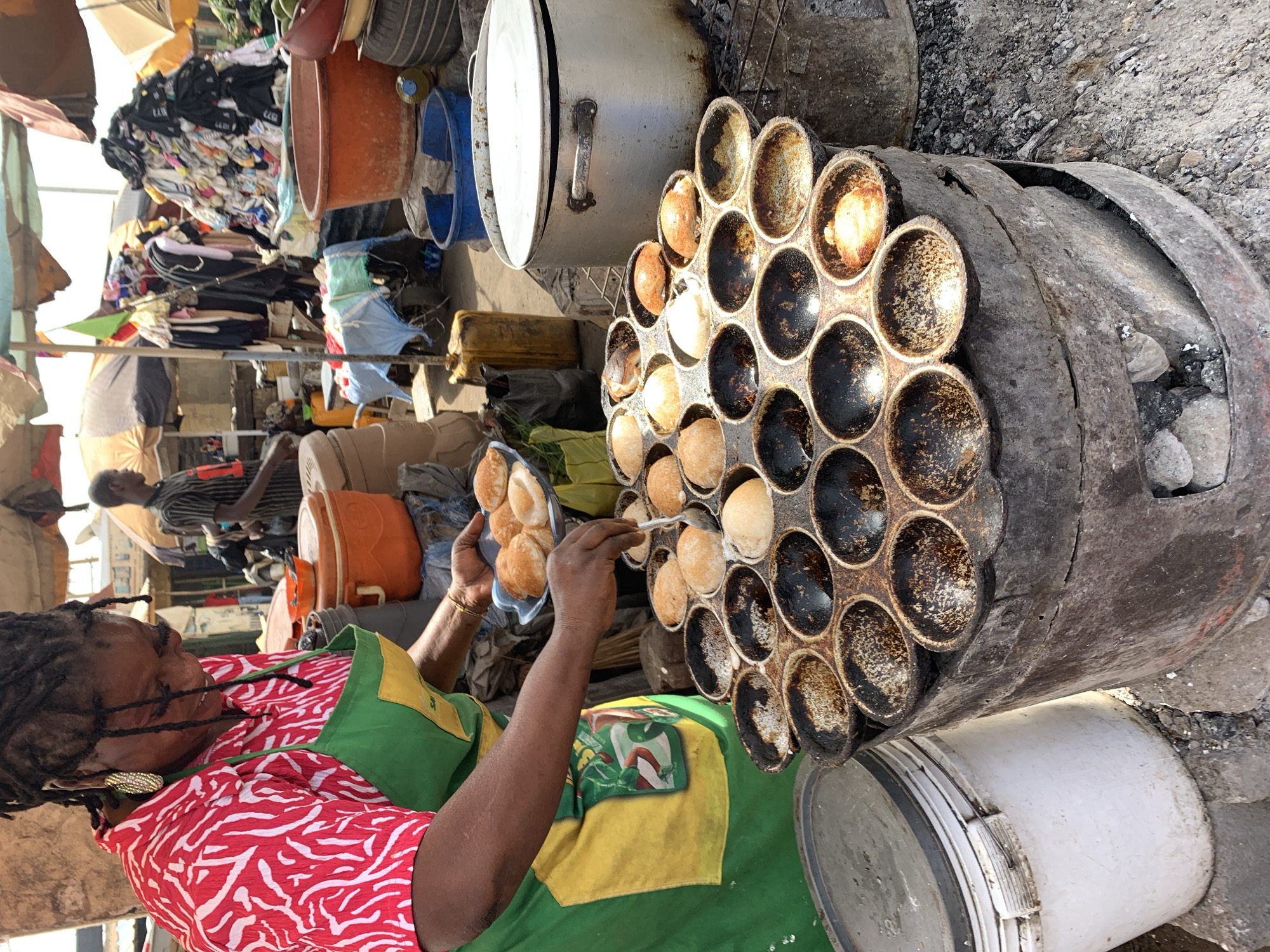 In Rafin Par, Nigeria, a street vendor wearing a red and white patterned shirt and a green and yellow apron is using a utensil to remove small, round, baked goods from a large, circular, multi-compartment cooking surface. The cooking surface is placed on a metal container that appears to be heated by coals or another heat source inside. The baked goods are being placed onto a clear plastic tray. In the background, there are piles of clothing and various buckets and containers, suggesting a market setting. Other people are visible in the background, going about their business.